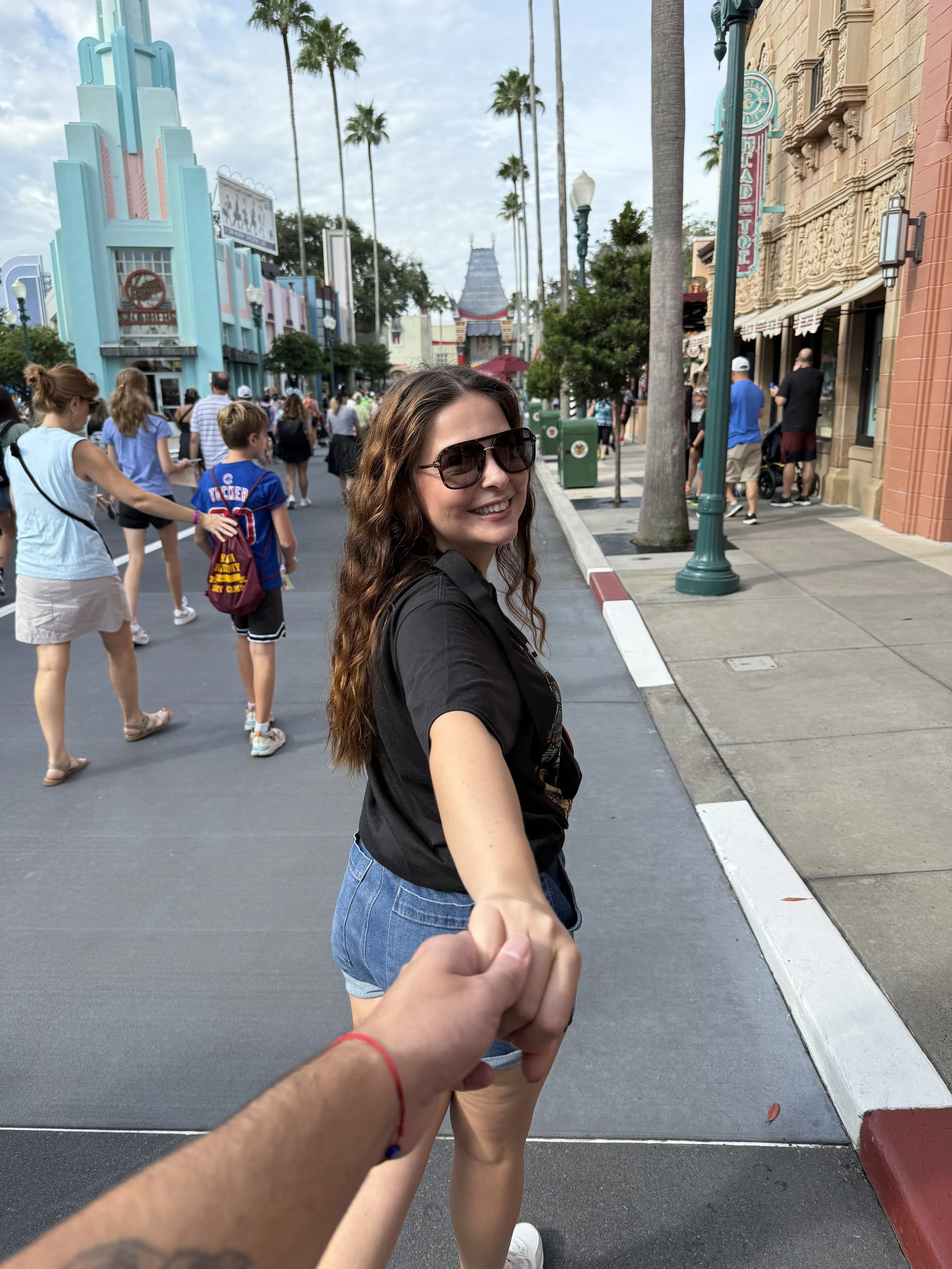 A woman wearing sunglasses and a black T-shirt holding hands with husband at Hollywood Studios. The background shows colorful buildings, palm trees, and other visitors walking around.
