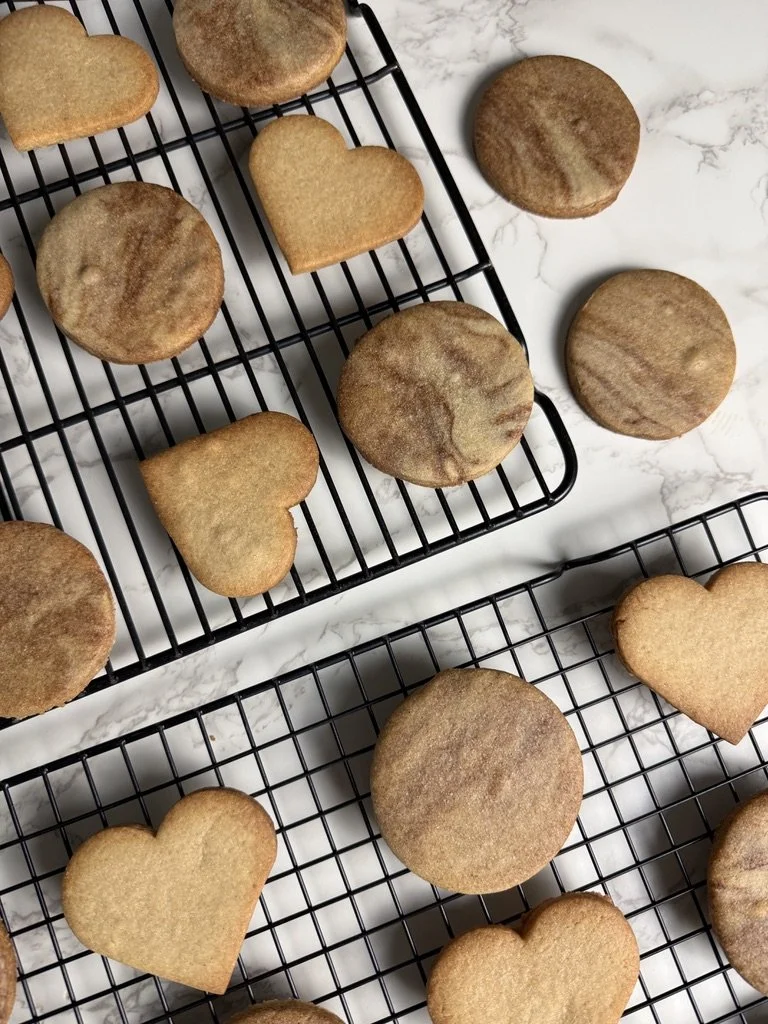 Assorted heart and round cookies cooling on black wire racks on a white marble surface.