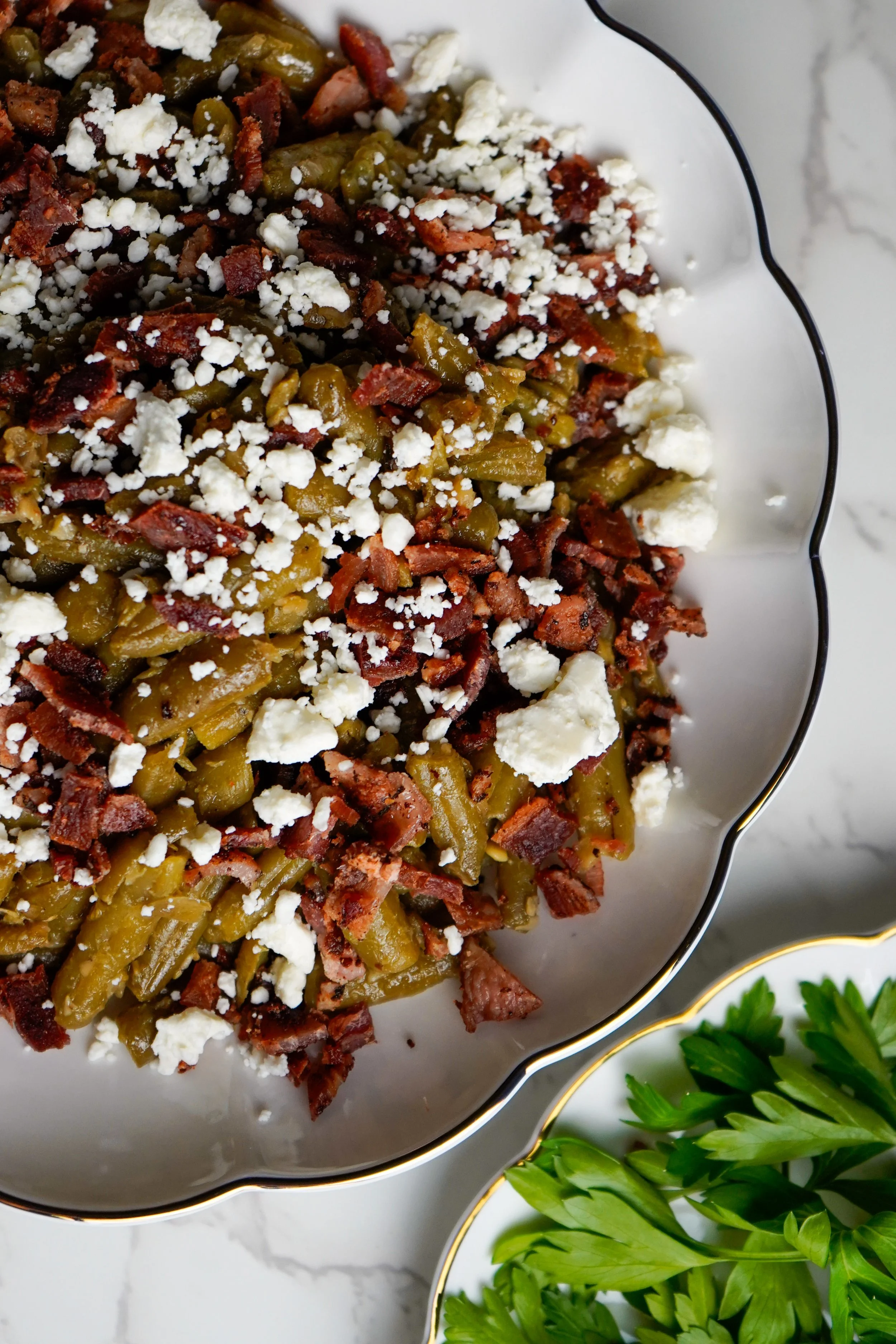 Baked green beans with bacon, crumbled feta cheese, and herbs in a white dish with black trim.