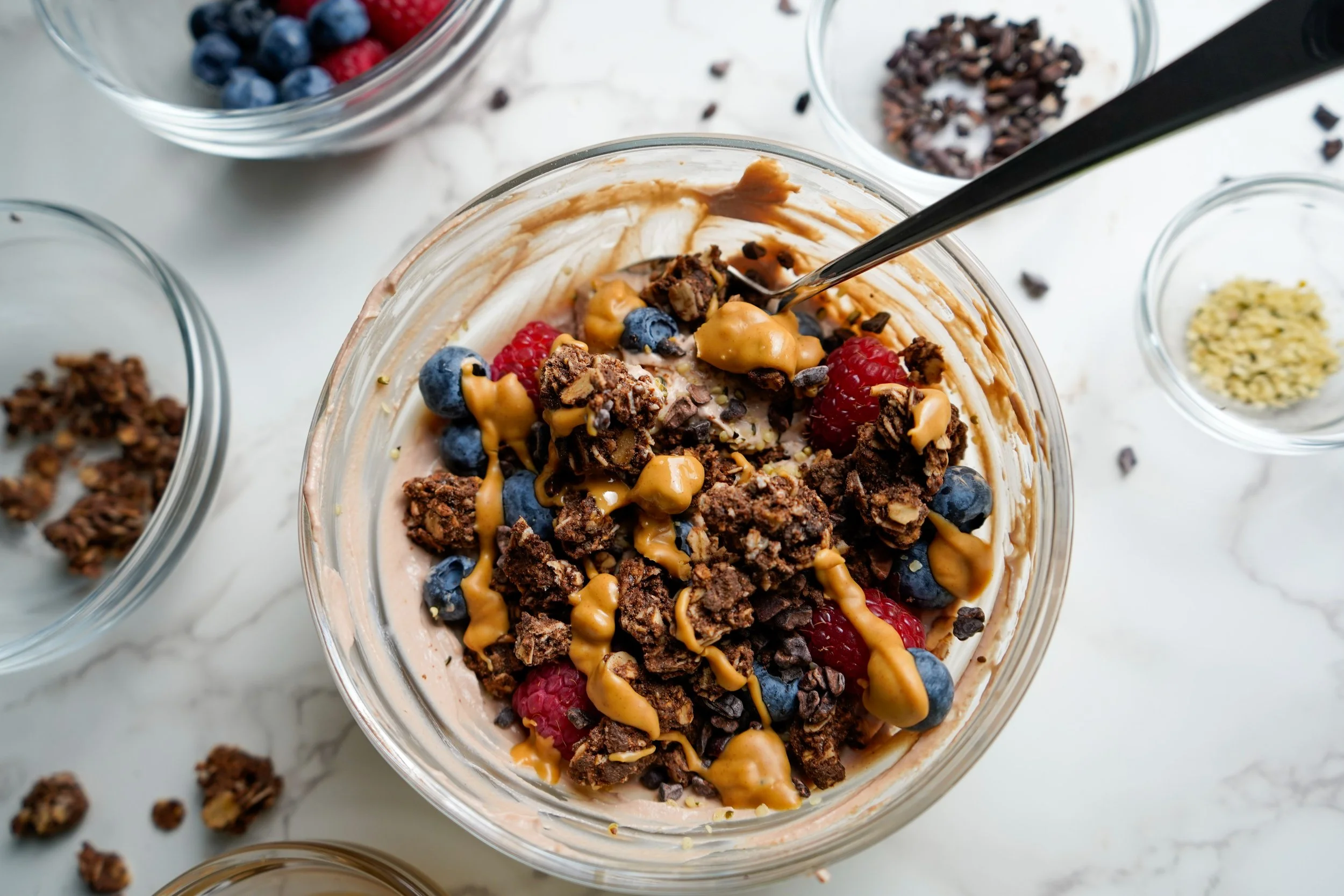 A glass bowl of protein yogurt topped with berries, granola, peanut butter, and cacao nibs, with small bowls of toppings on a white surface.