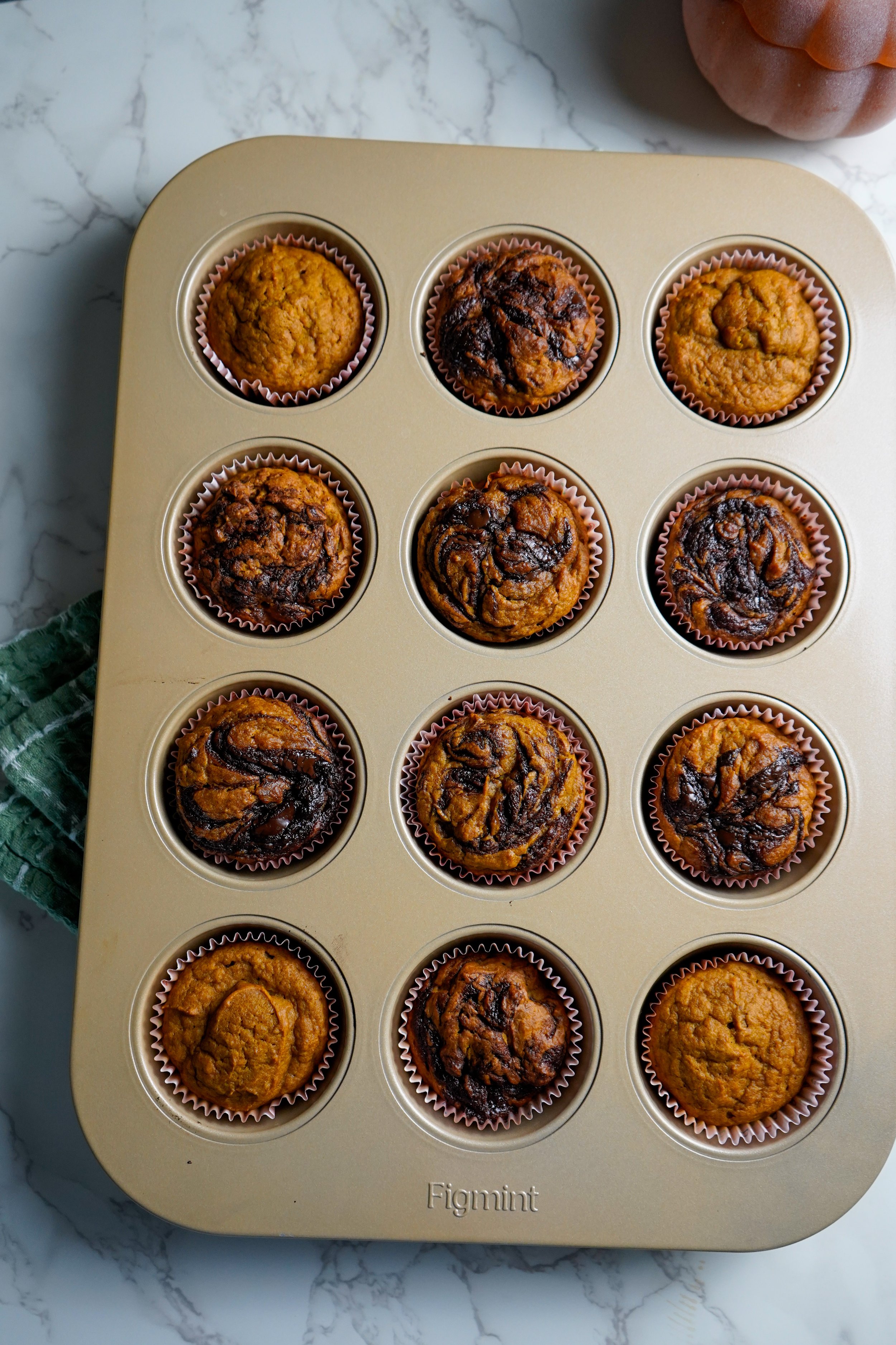 A metal muffin tin filled with twelve muffins, some with chocolate swirls and others with a plain or slightly golden appearance, on a white marble surface.