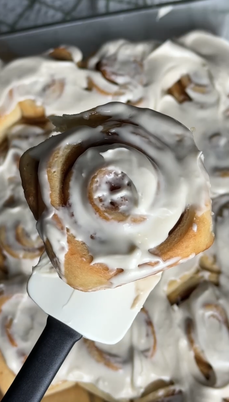 Close-up of a cinnamon roll with white icing, held with a spatula in front of a tray of similar cinnamon rolls with icing.