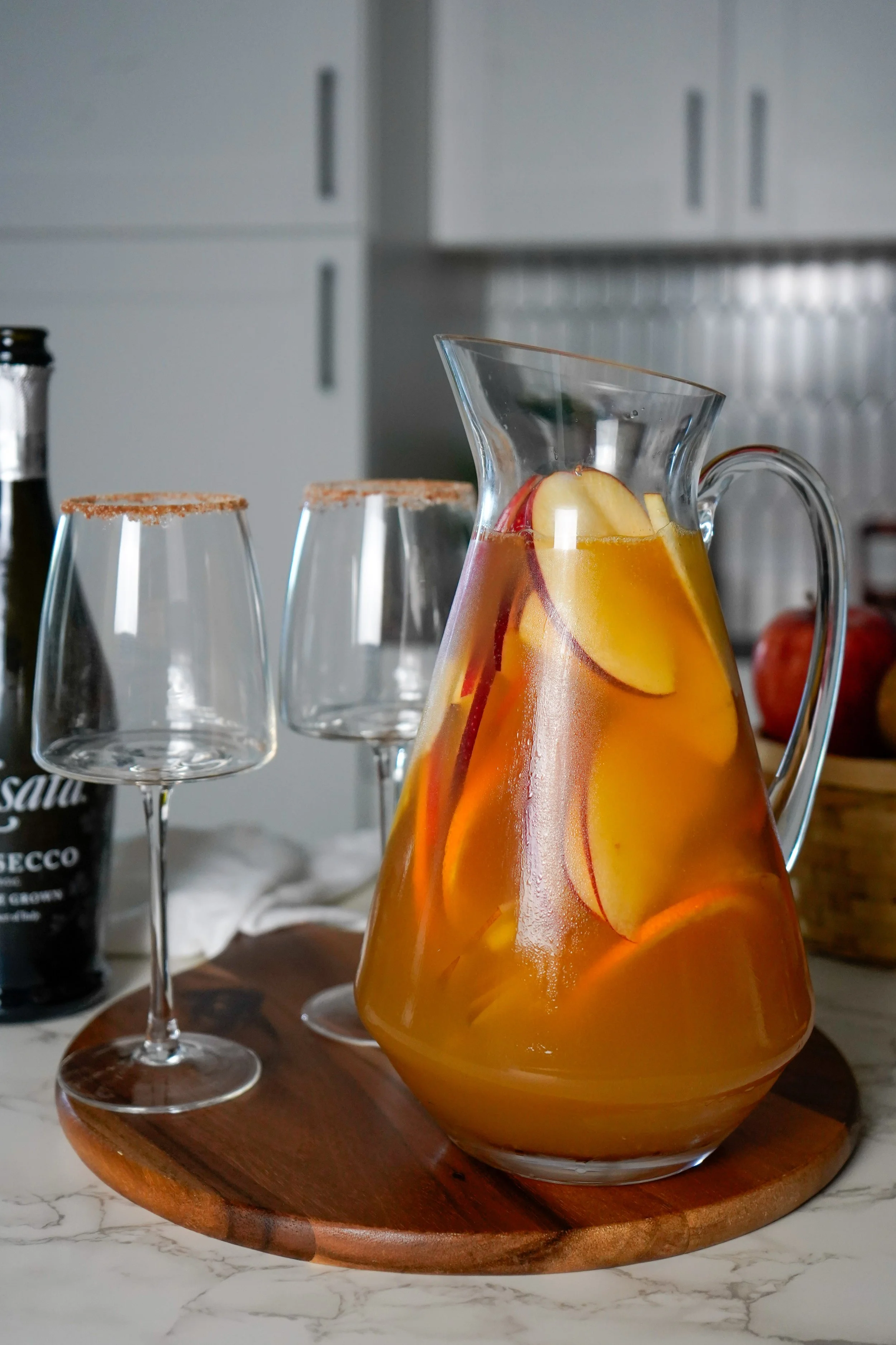 A glass pitcher filled with sliced apples , pears, and orange along with cocktail liquid, set on a wooden tray; two empty wine glasses with cinnamon-sugar rims beside it, on a kitchen countertop.