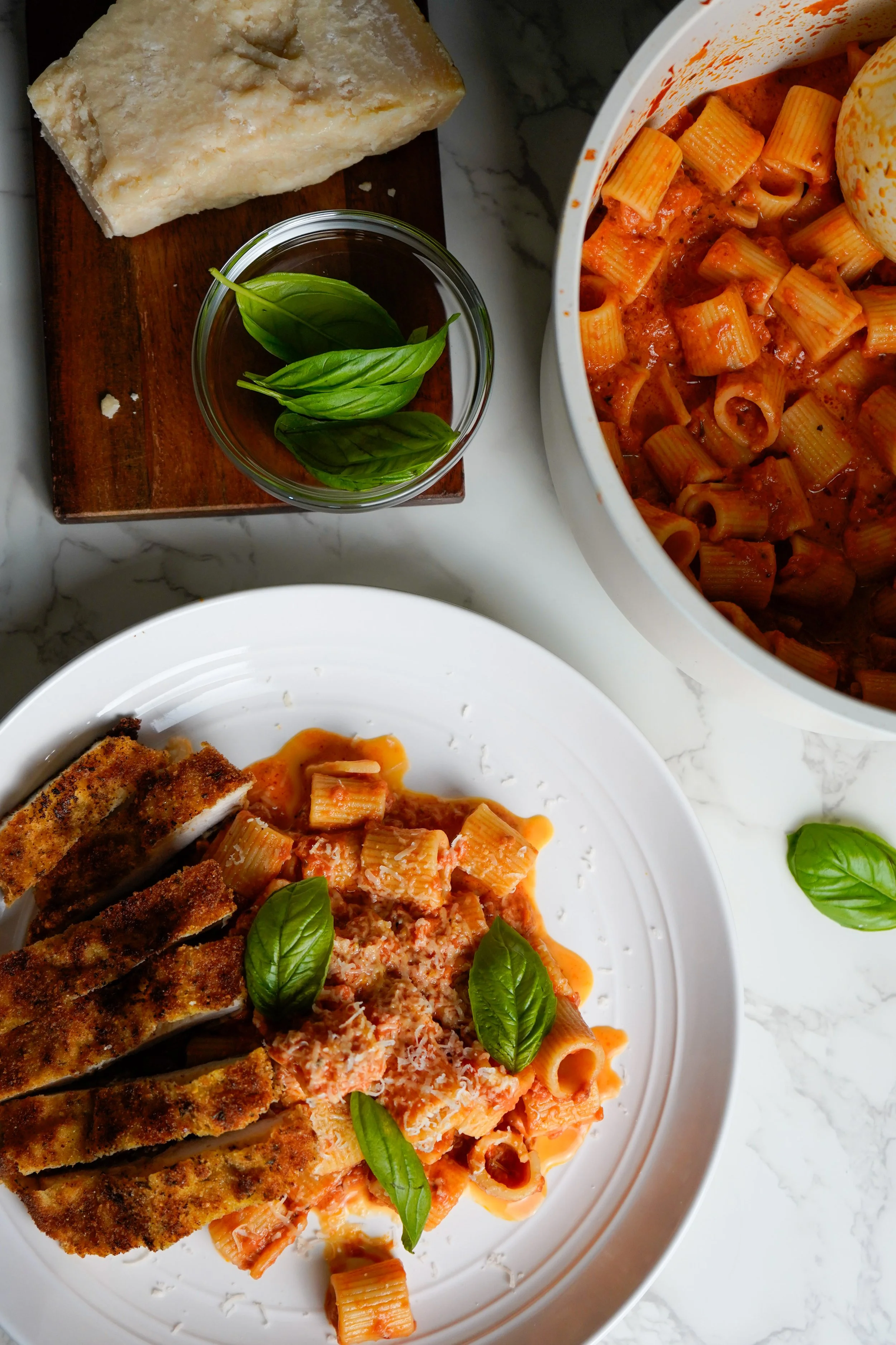 Plate of cooked Mezzi rigatoni pasta with tomato sauce, topped with grated cheese and fresh basil leaves, served with breaded and fried meat cutlets; also includes a bowl of more pasta and basil leaves on a marble surface.