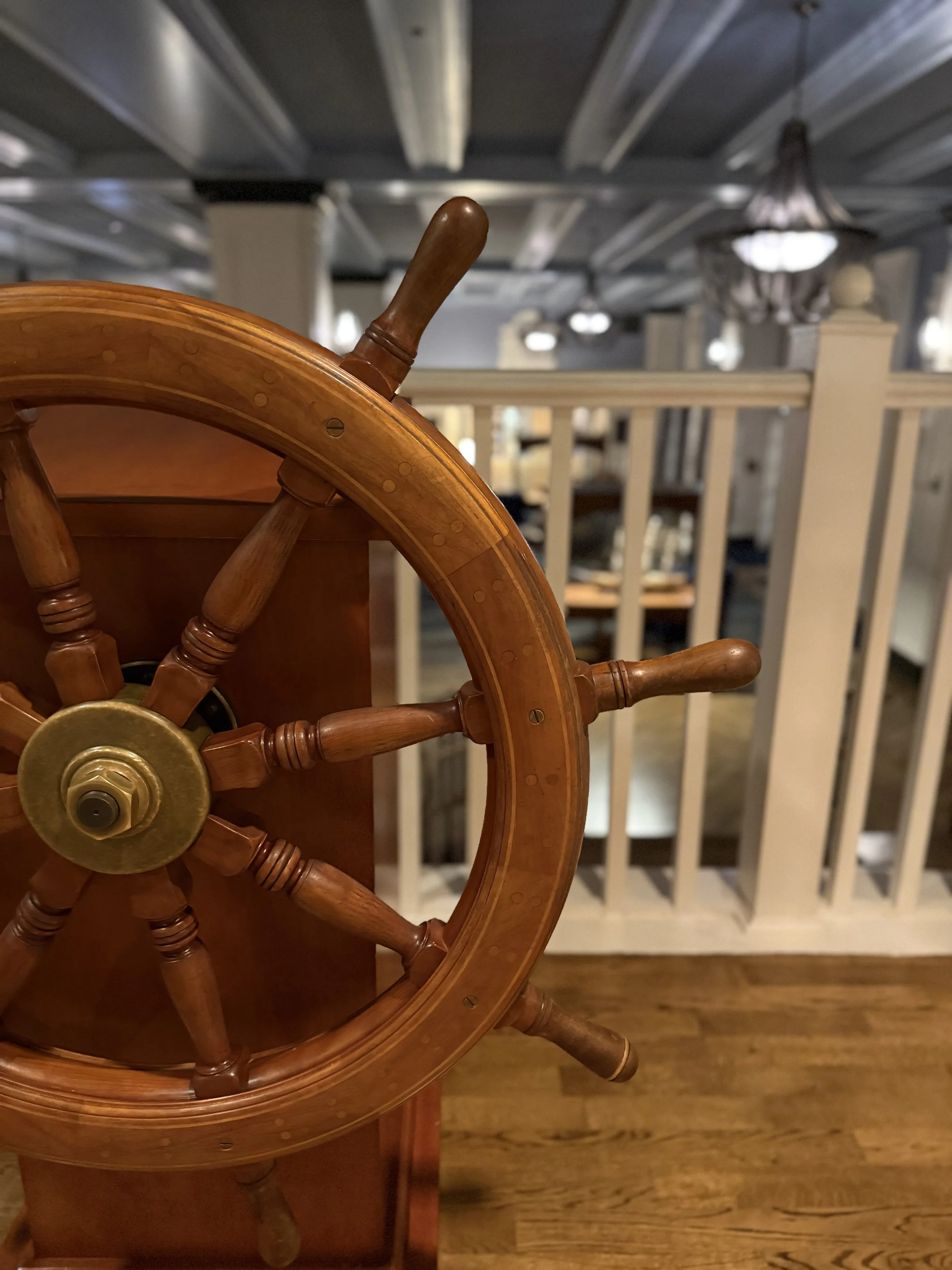Close-up of a wooden ship's steering wheel on a wooden table in a room with a railing and hanging light fixtures.