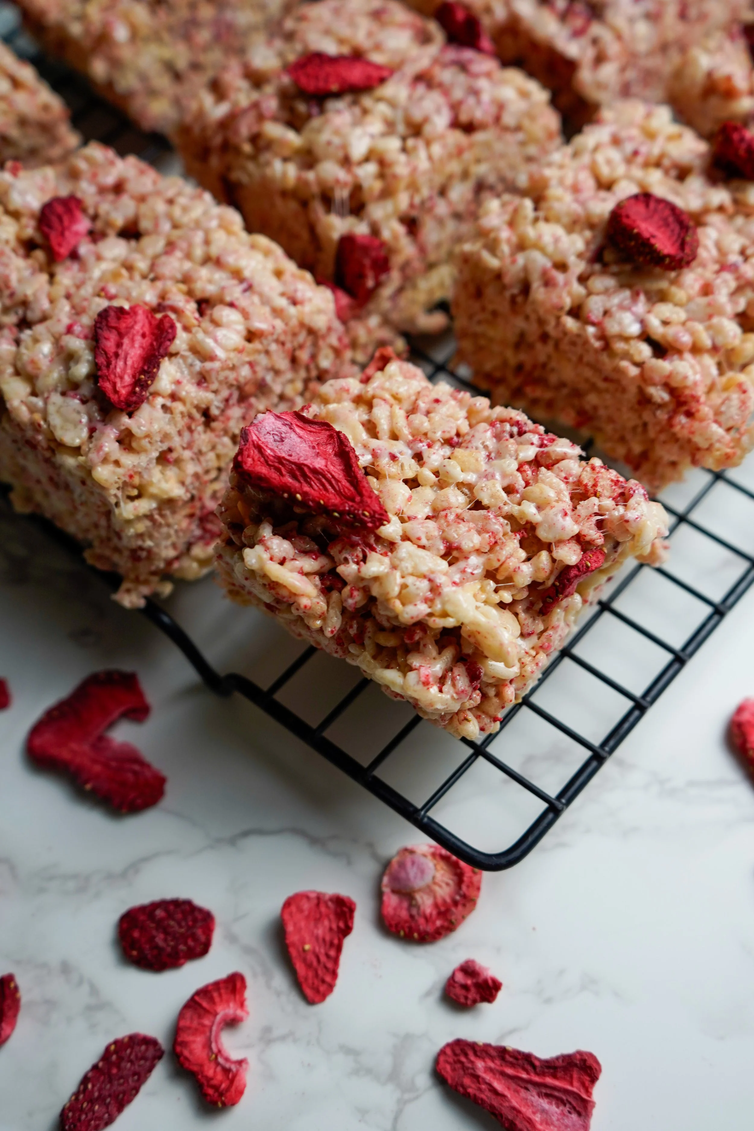Rice crispy treats topped with dried strawberries, set on a black wire cooling rack on a marble surface with additional dried strawberries scattered around.