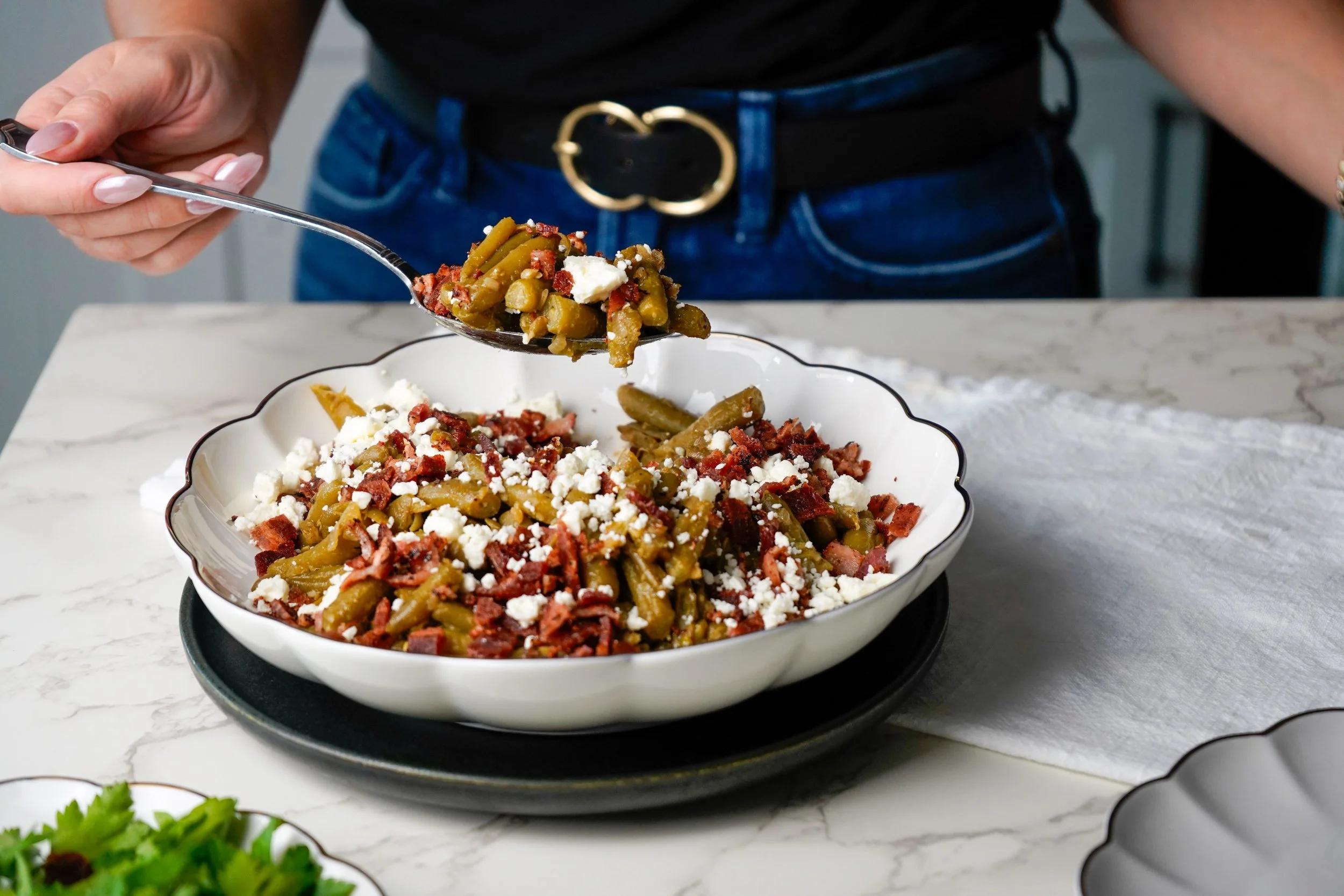Person serving a dish of green beans, bacon, and crumbled cheese from a spoon into a bowl on a kitchen counter.