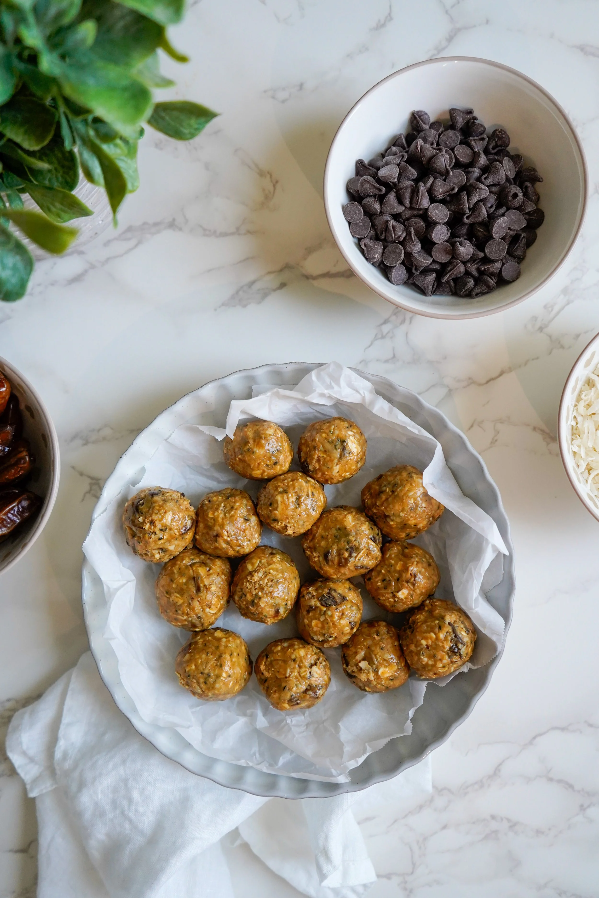 Homemade energy balls on a plate lined with parchment paper, surrounded by bowls of chocolate chips, dates, and shredded coconut on a marble counter.