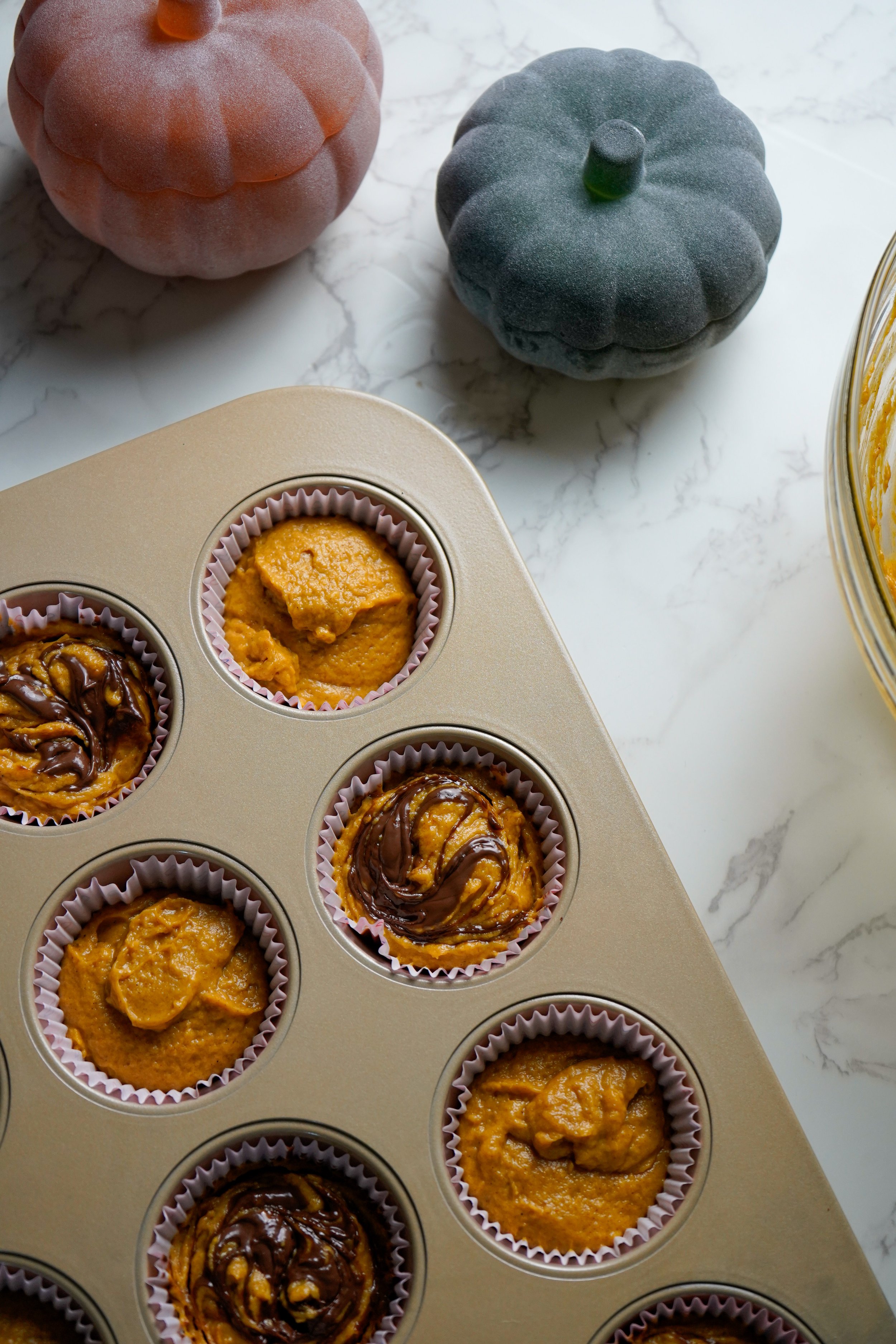 Unbaked muffins in a metal pan with pumpkin-shaped decor in the background, one orange and one green, on a white marble countertop.
