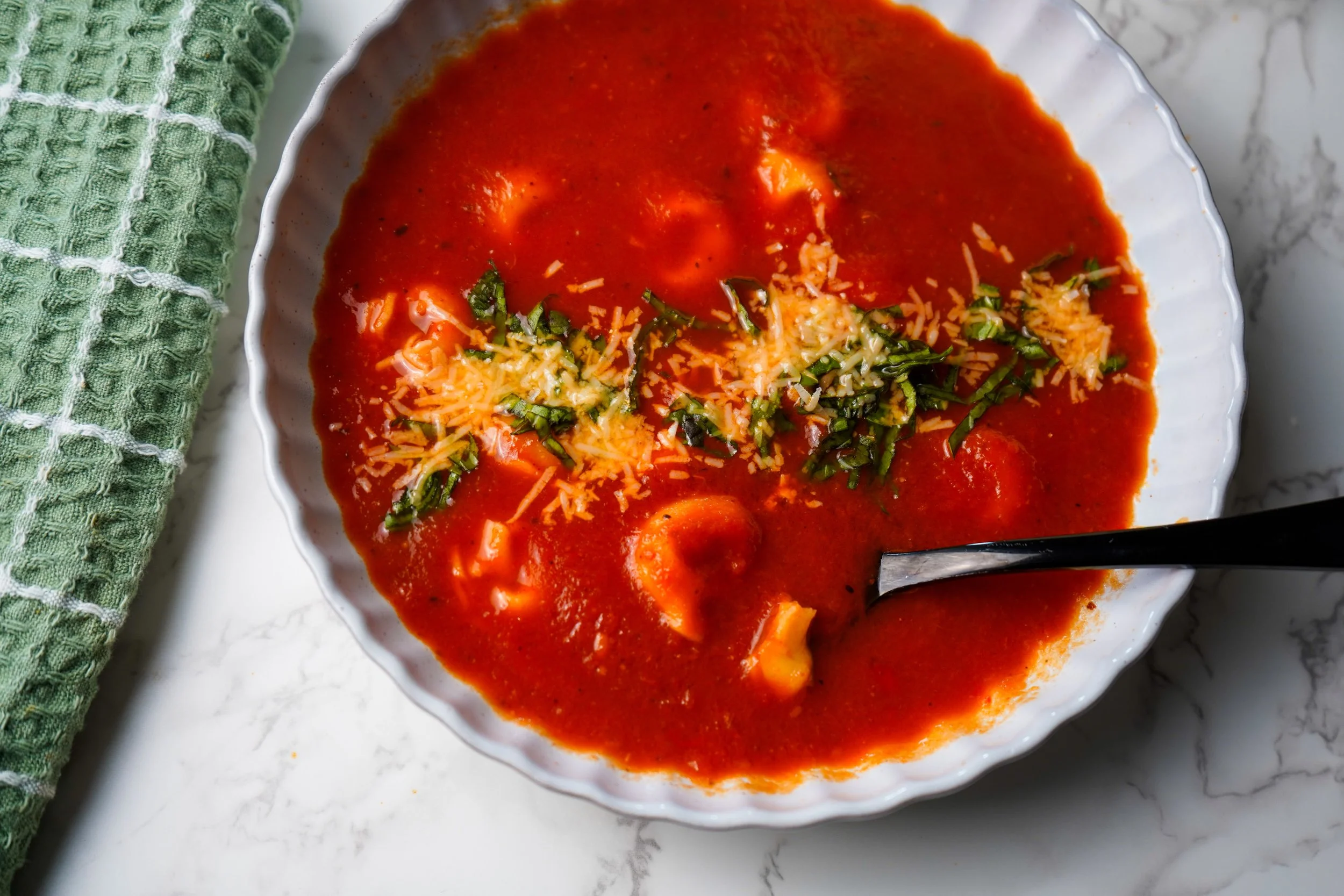 Bowl of tomato soup garnished with shredded cheese and chopped herbs, on a white marble surface with a green towel nearby.