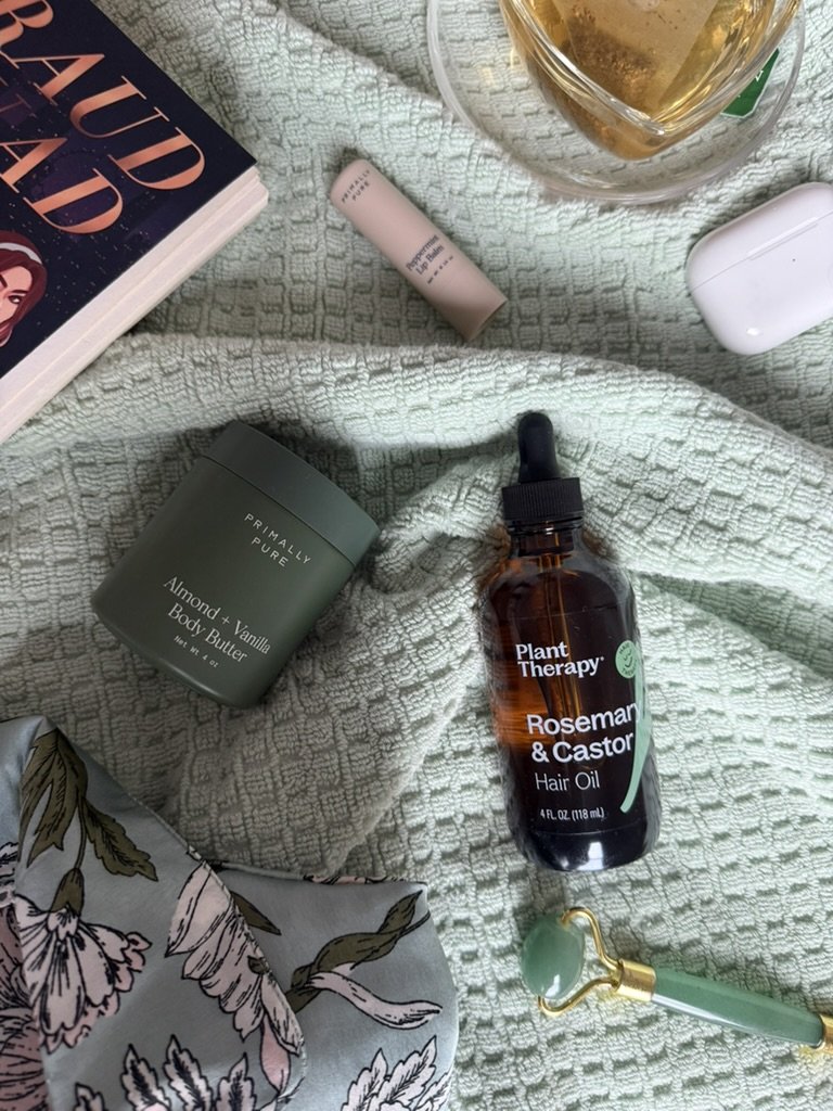 A flat lay of various personal and beauty items on a textured, light green blanket. Includes a book a tube of lip balm, a jar of almond vanilla body butter, a bottle of Plant Therapy rosemary and castor hair oil, a pair of white wireless earbuds, part of a floral-patterned fabric, and a jade facial roller.