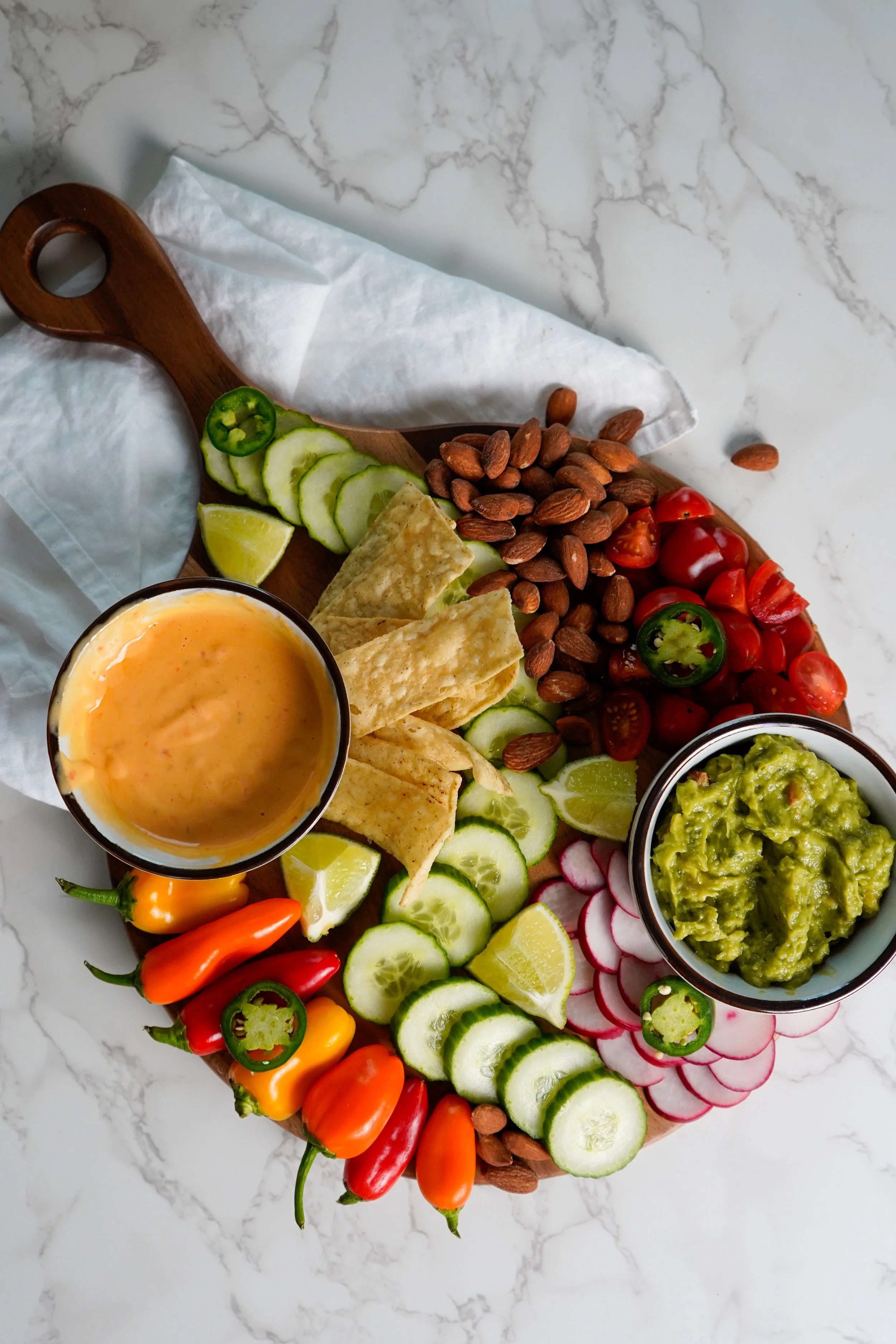 Colorful charcuterie board with sliced cucumbers, radishes, cherry tomatoes, jalapillos, yellow and orange peppers, lime wedges, tortilla chips, and two bowls of dips on a white marble surface.