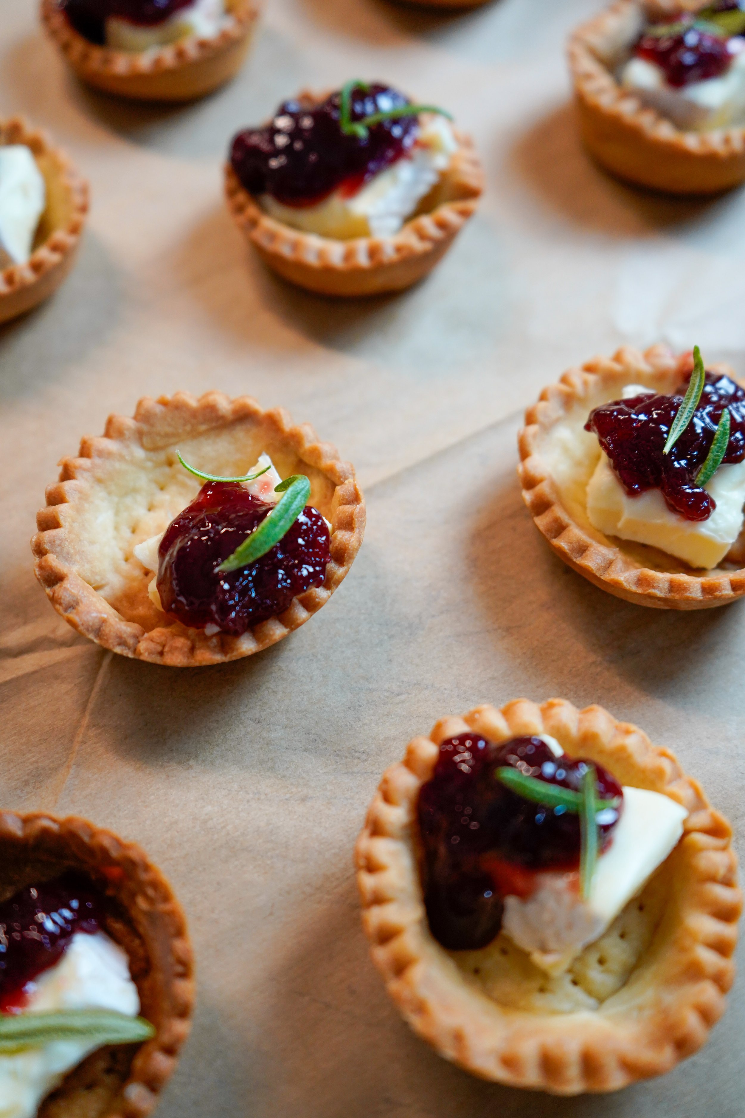 Mini tartlets filled with cheese and topped with sour cherry compote and rosemary sprigs, arranged on parchment paper.