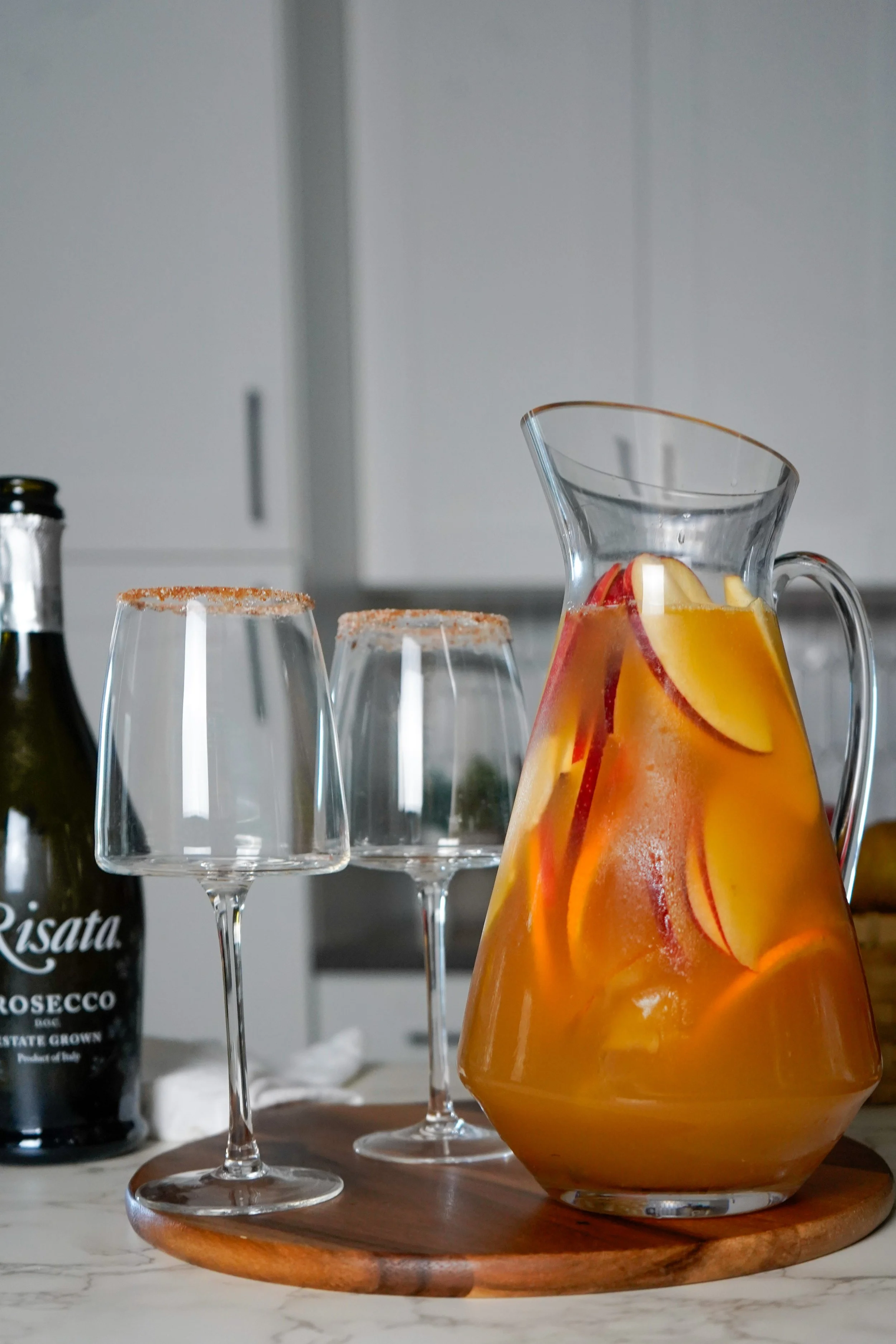 A glass pitcher filled with apple cider sangria and slices of apple, pear, and oranges alongside two empty wine glasses with cinnamon sugar-rimmed edges, and a bottle of Risata Prosecco on a wooden serving board on a kitchen countertop.