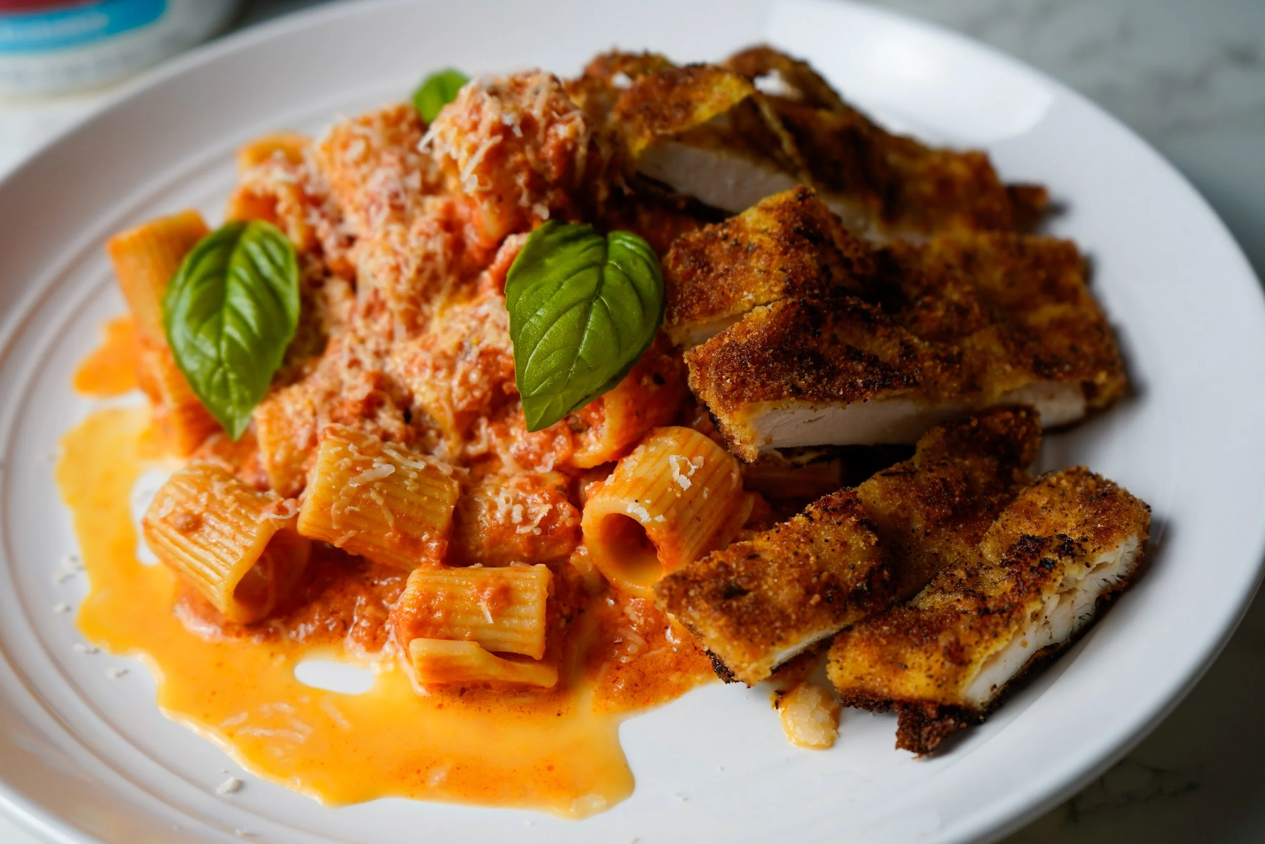 A plate of Italian food featuring Mezzi rigatoni pasta with spicy tomato sauce, breaded and fried chicken cutlets, garnished with basil leaves and grated parmesan cheese.