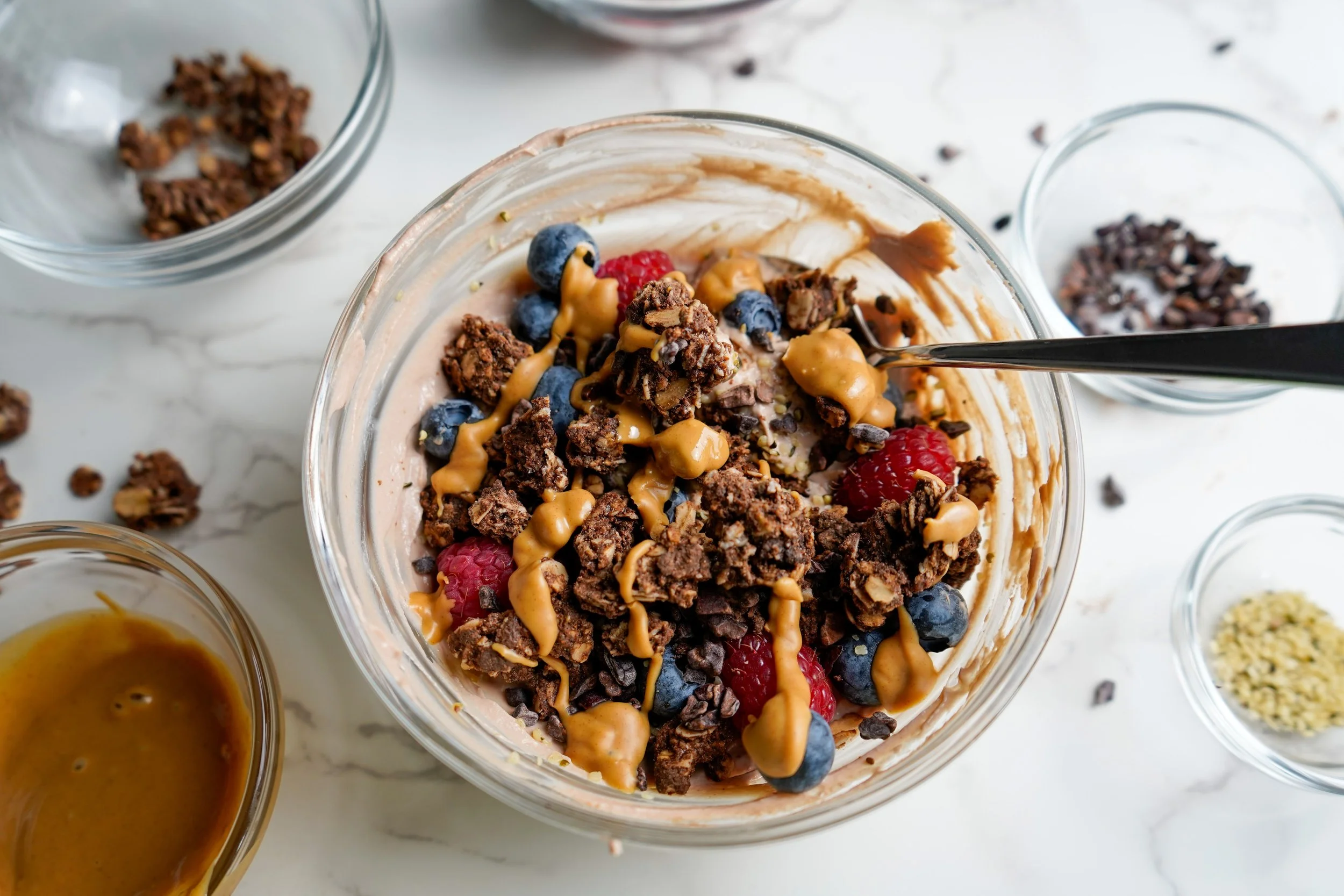 A bowl of protein yogurt topped with cocoa nibs, blueberries, raspberries, and peanut butter drizzle, with additional ingredients and bowls surrounding it on a marble surface.