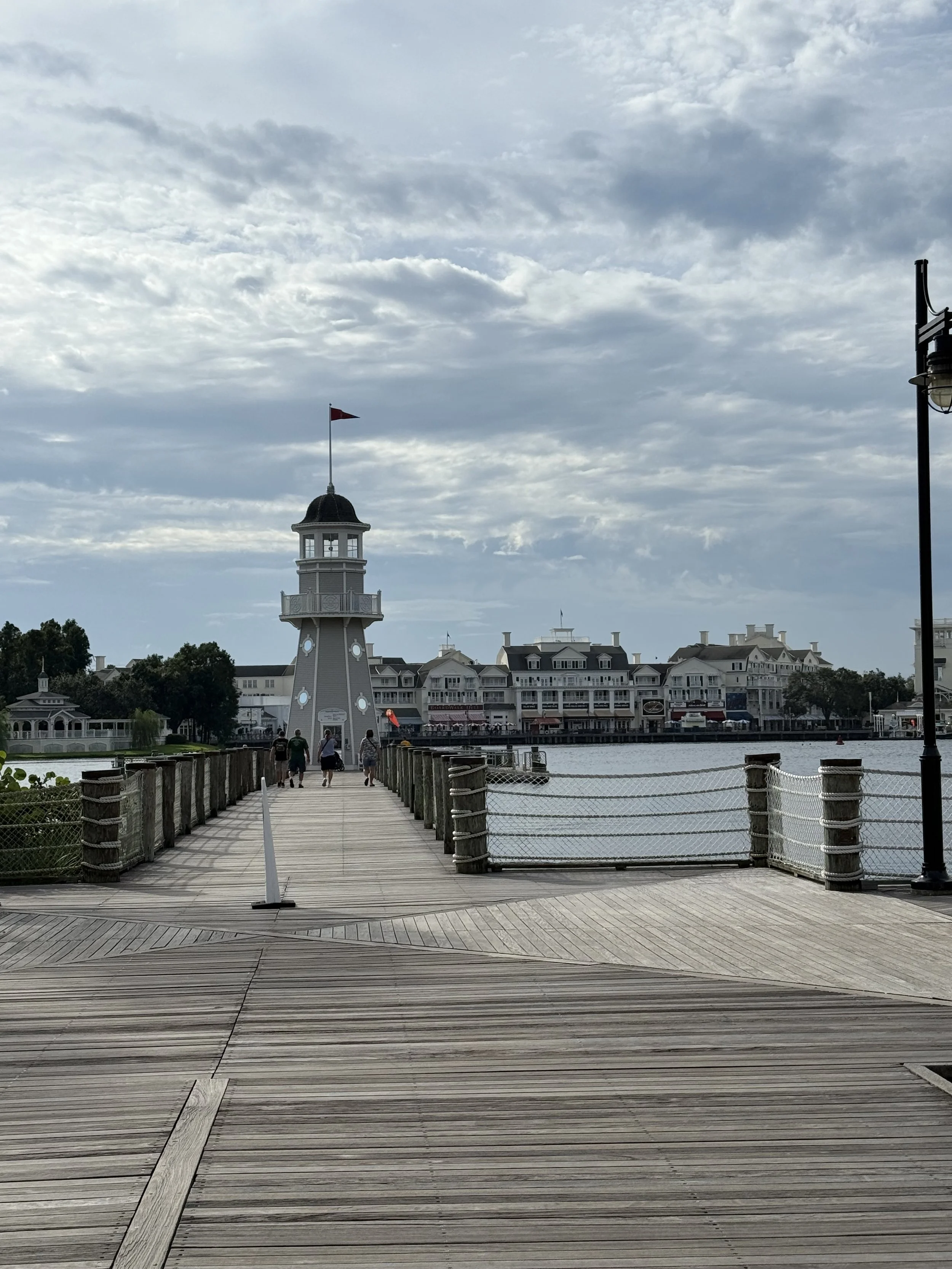 A wooden pier leading to a lighthouse with a red flag, overlooking water with buildings in the background, under a partly cloudy sky.