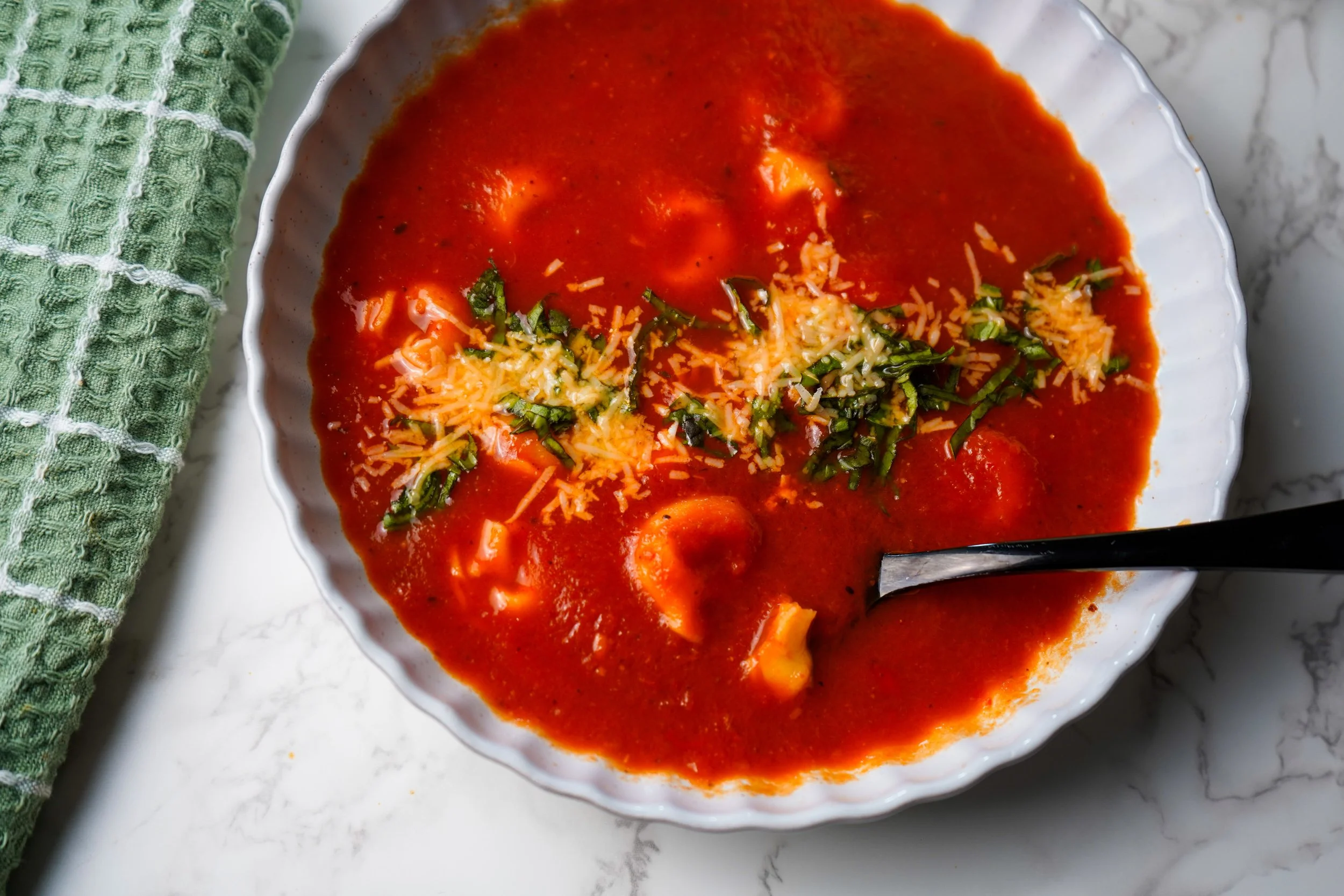 Bowl of tomato soup garnished with shredded cheese and chopped herbs on a white marble surface.