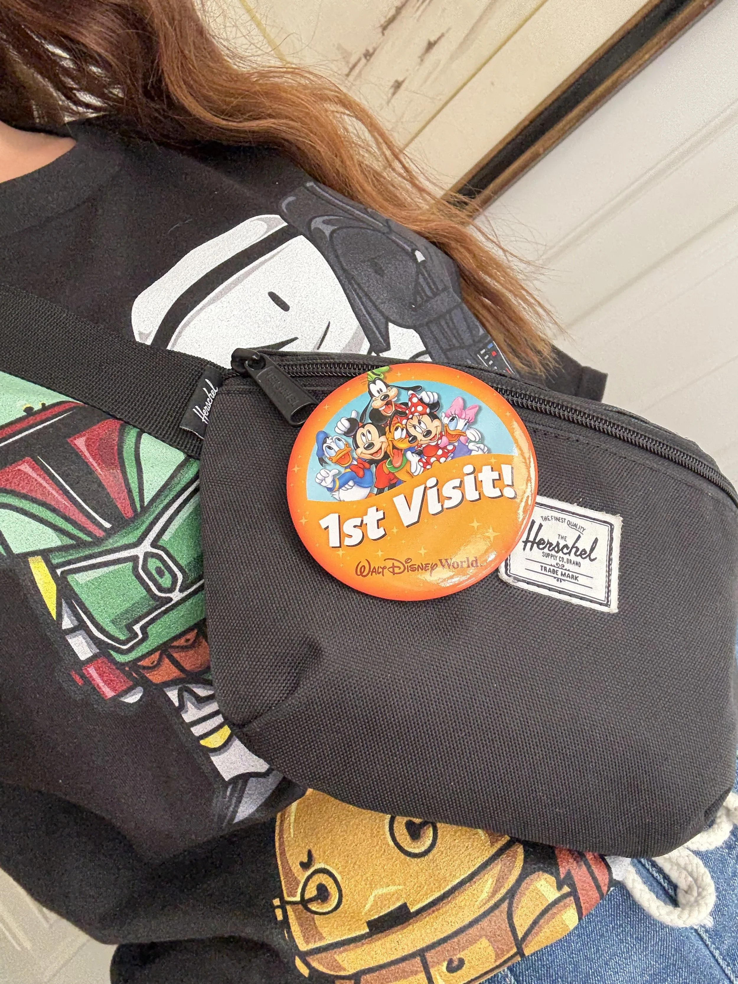 Close-up of a woman wearing a black The Mandalorian t-shirt with a graphic print, carrying a black Herschel bag with a Disney World badge that reads '1st Visit!' featuring Disney characters Mickey Mouse, Minnie Mouse, Donald Duck, Daisy Duck, Goofy, and Pluto, attached to the bag.
