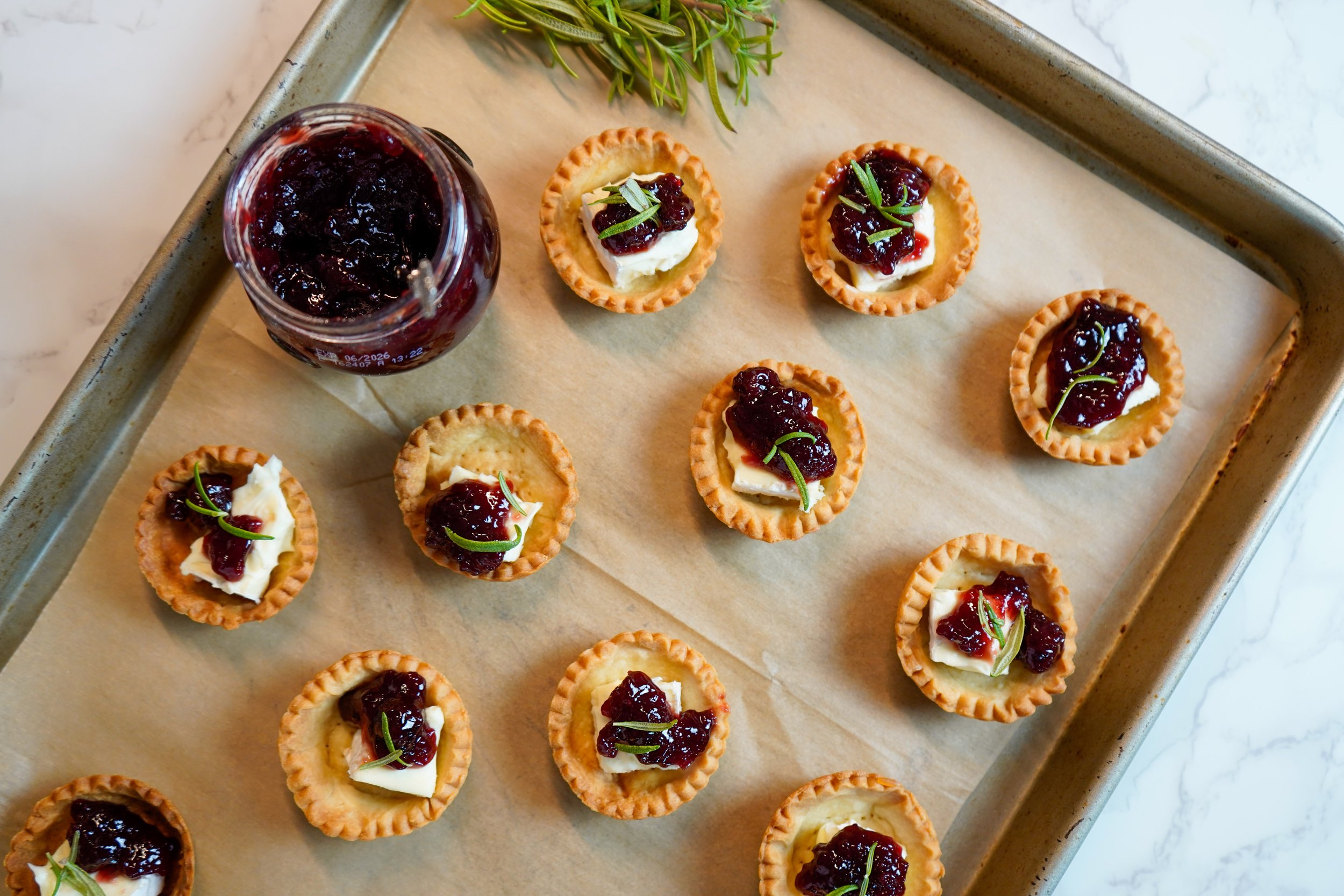 Mini tartlets topped with Saint Angel cheese, sour cherry jam, and small green herbs, arranged on a parchment-lined baking sheet with a jar of berry jam and some herbs nearby.