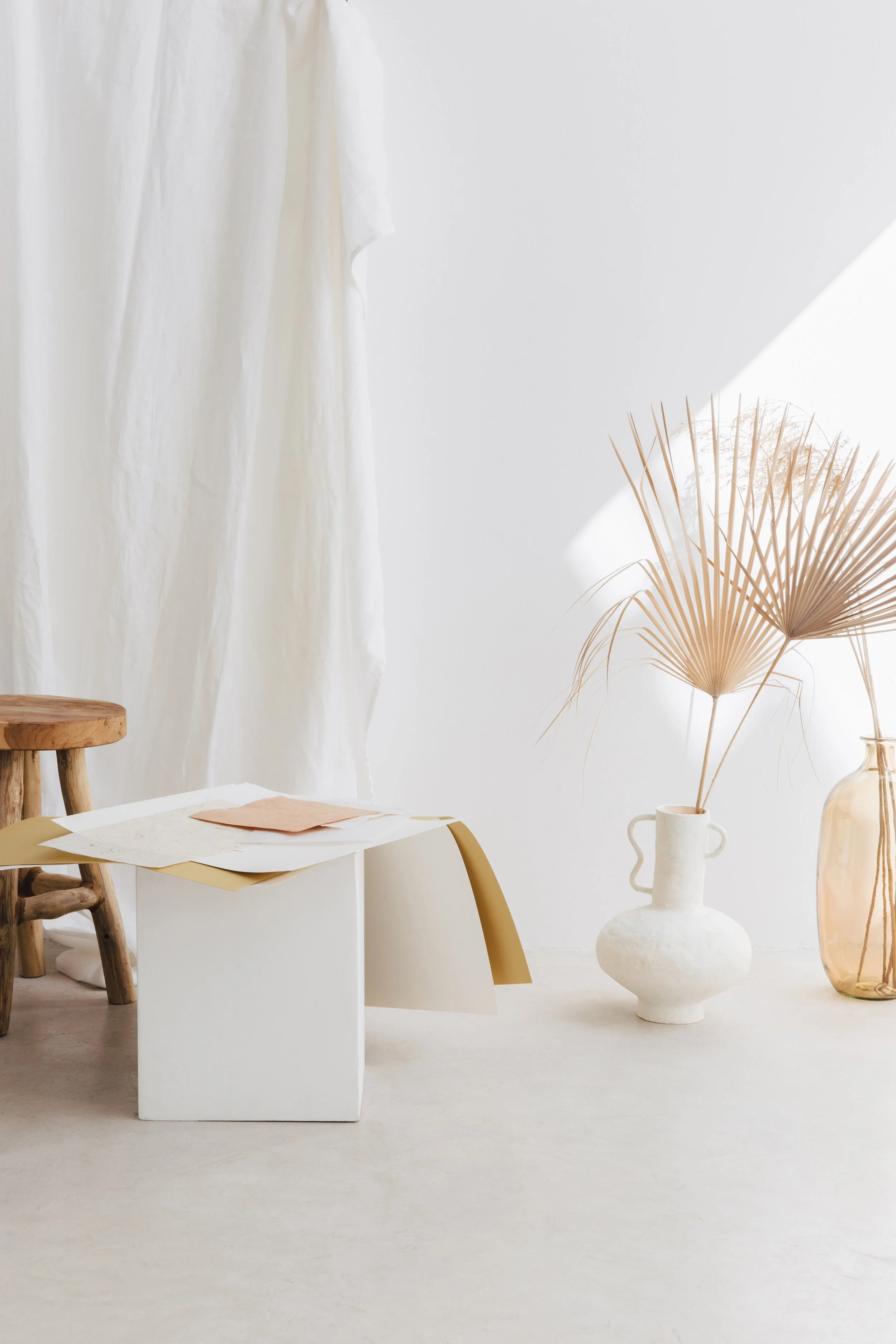 Minimalist interior with a white fabric drape, a small wooden stool, beige and white papers on a modern white table, and a ceramic vase with dried palm leaves, set against a white wall with sunlight.
