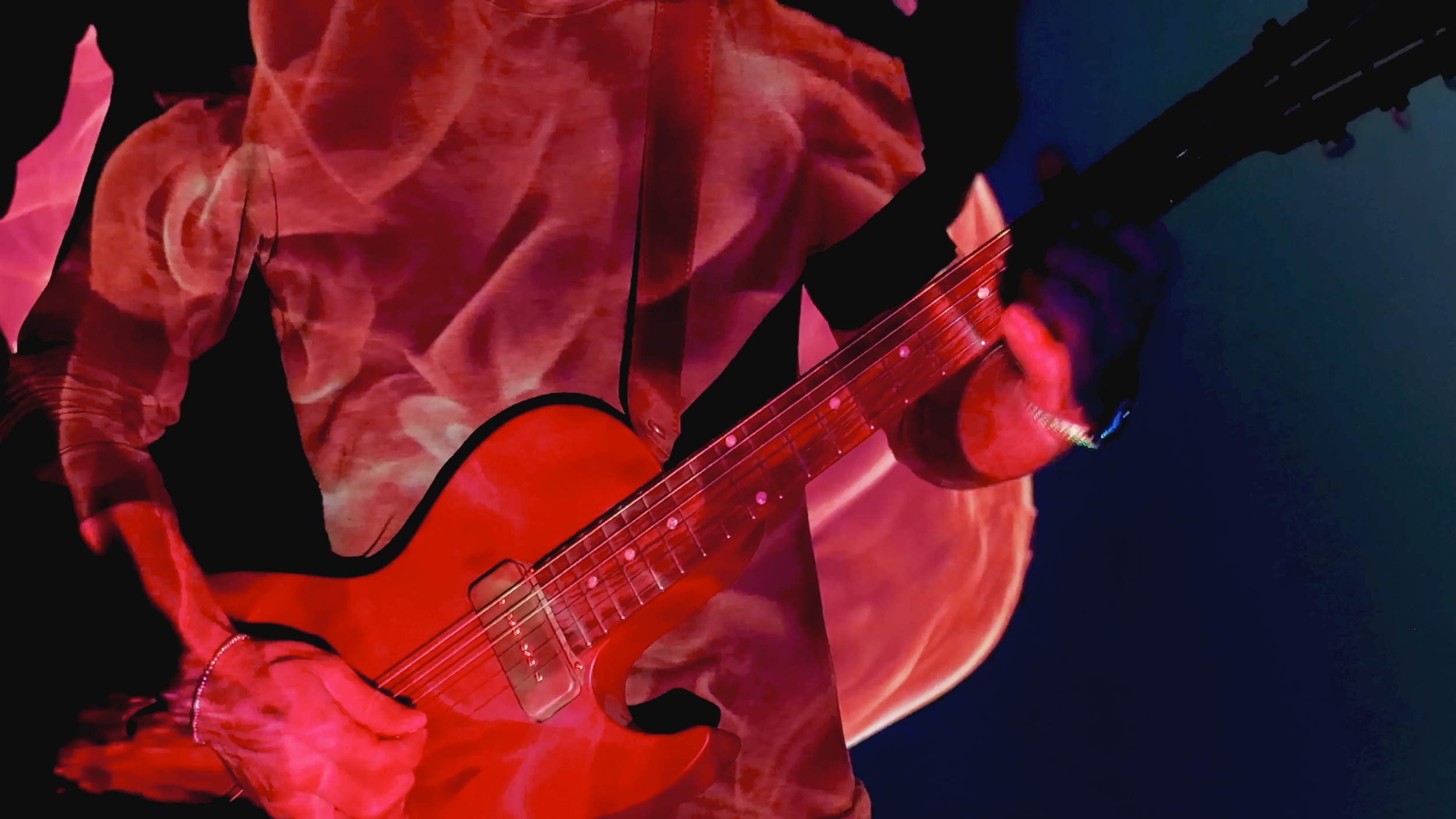 Close-up of a person playing an orange electric guitar, wearing a red tie-dye hoodie against a dark background.