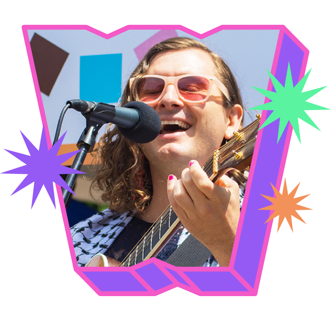 Evan Greer with shoulder length brown hair and pink sunglasses sings into a microphone while playing a guitar