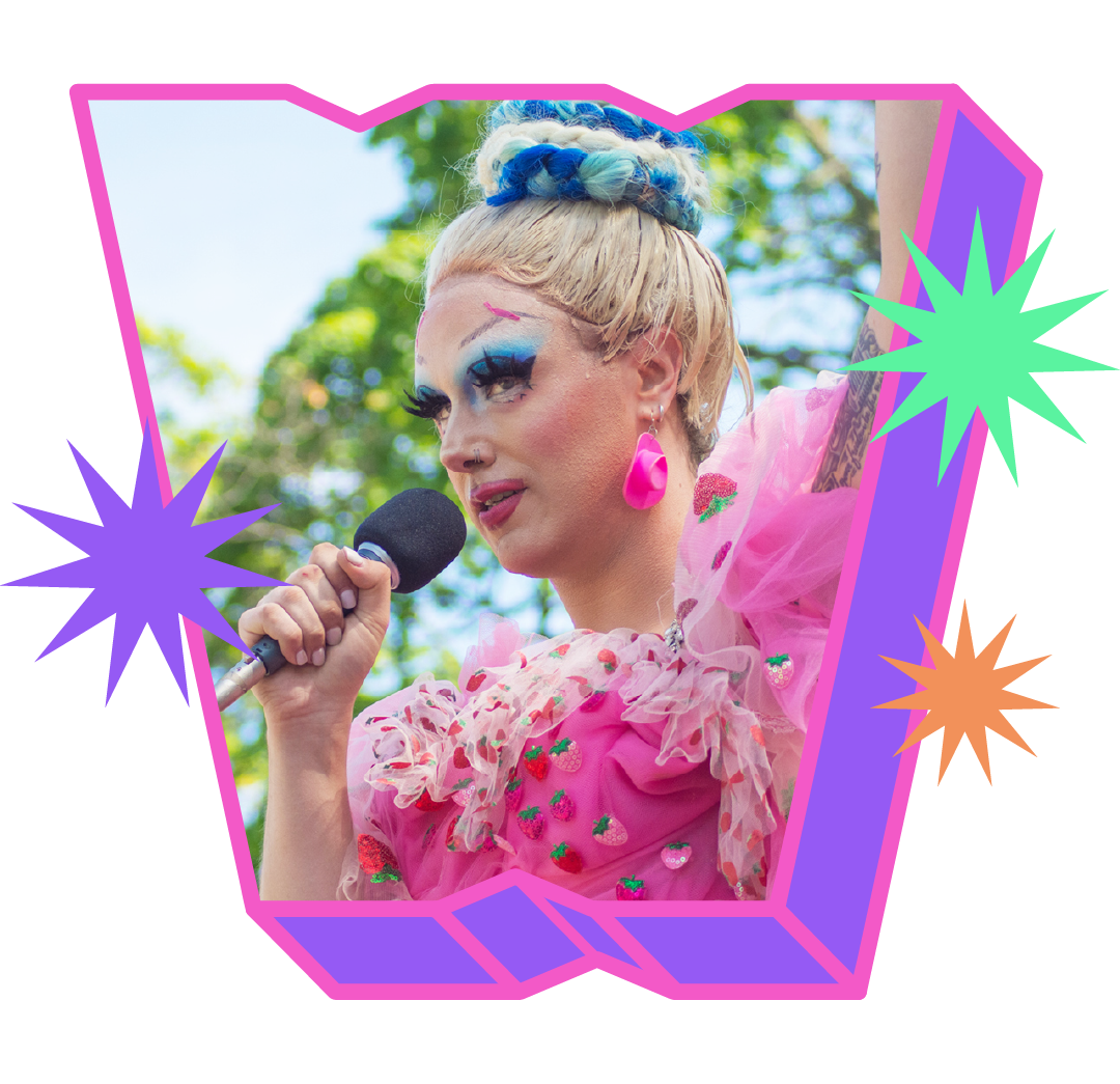 Missy Steak, a drag queen in a pink frilly dress, speaks into a microphone and holds a fist in the air