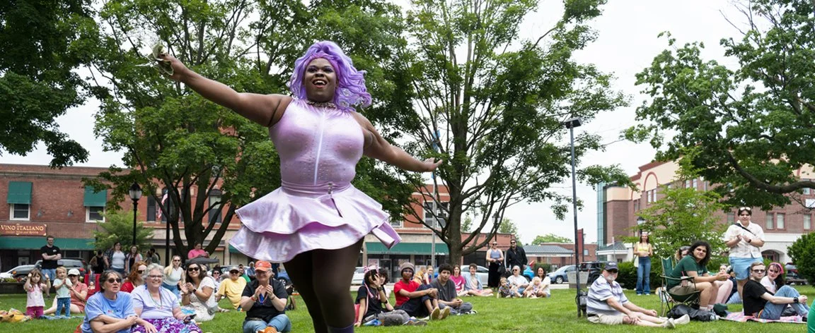 A drag queen with purple hair and a purple tutu dances through a crowd of people sitting on a green lawn