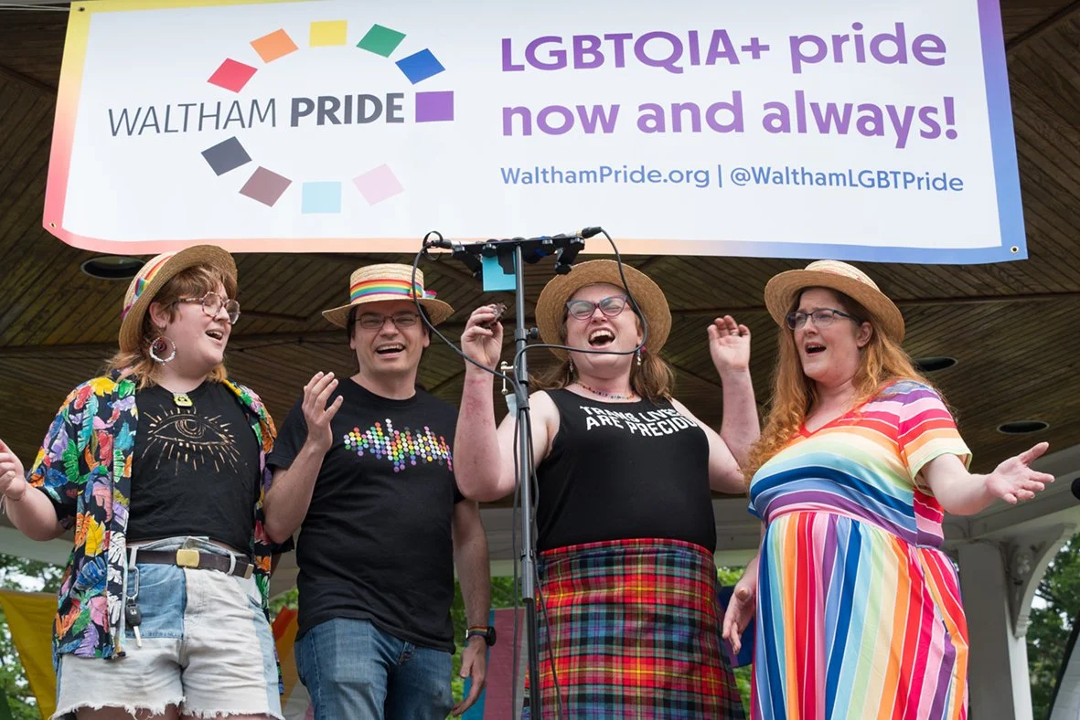Four people in colorful outfits and wearing straw boater hats singing into a microphone in front of a Waltham Pride "LGBTQIA+ pride now and always!" banner