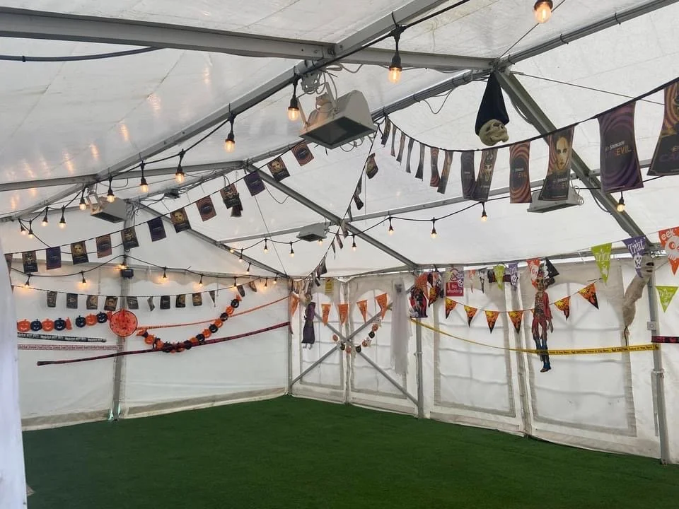 Decorated tent with Halloween and autumn-themed banners, string lights, and hanging decorations including paper lanterns, masks, and paper cutouts. The floor is covered with green artificial grass.
