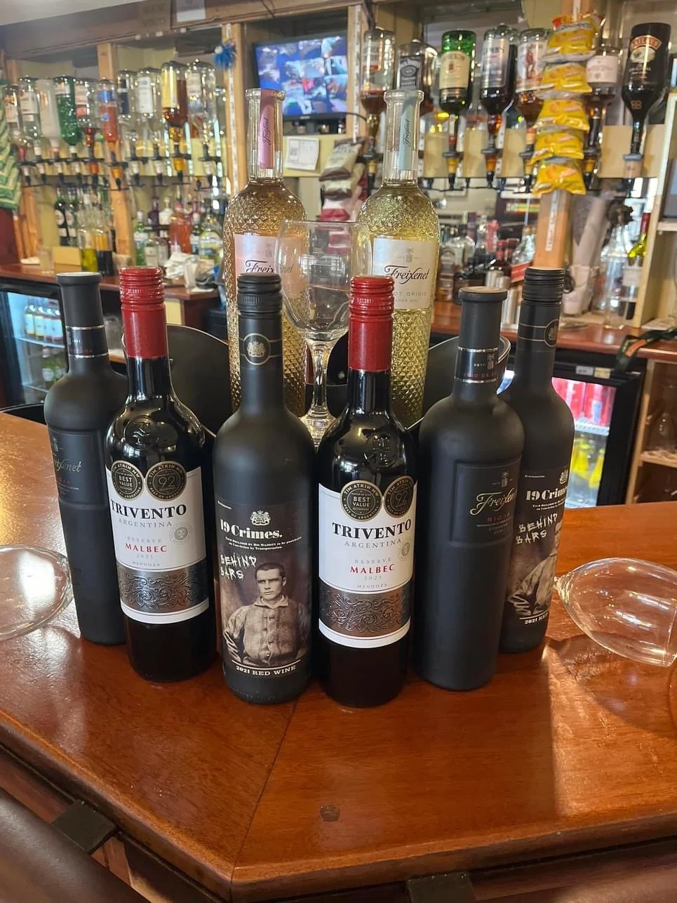 Six wine bottles on a wooden bar counter with a background of liquor bottles and a TV screen. Two bottles have black labels, two have white labels, and two have decorative bottles with yellow liquid inside. There are also two wine glasses and a small