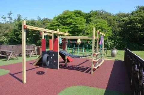 Children's playground with climbing structures, a tire swing, and a woven rope tunnel, surrounded by trees and outdoor furniture.