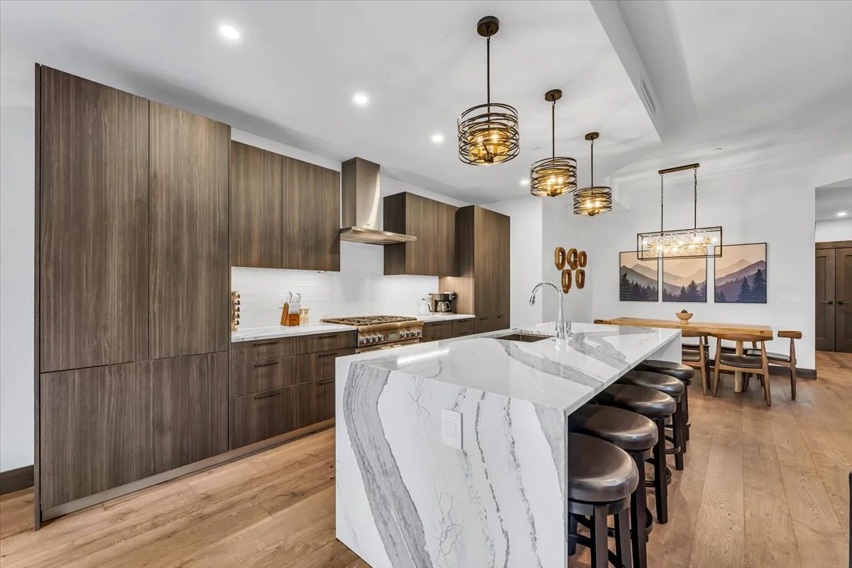 Contemporary kitchen with dark wood cabinetry, a marble island with bar stool seating for 5 people, and a dining area in the background.