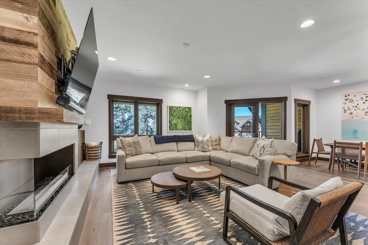 This cozy yet contemporary living area features a large seating area, warm stone fireplace, hardwood floors, an area rug and abundant natural light streaming through multiple windows. A patio door opens seamlessly to a private outdoor retreat.