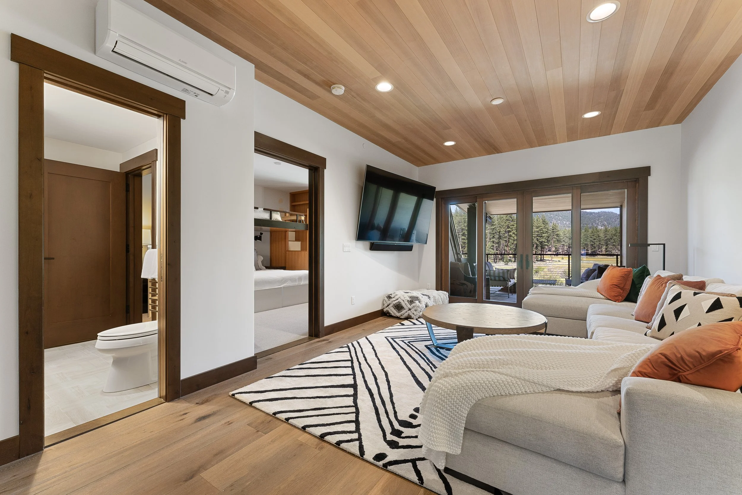 This cozy living space features natural wood ceiling, a large sofa with expansive windows framing serene forest views, and abundant natural light.