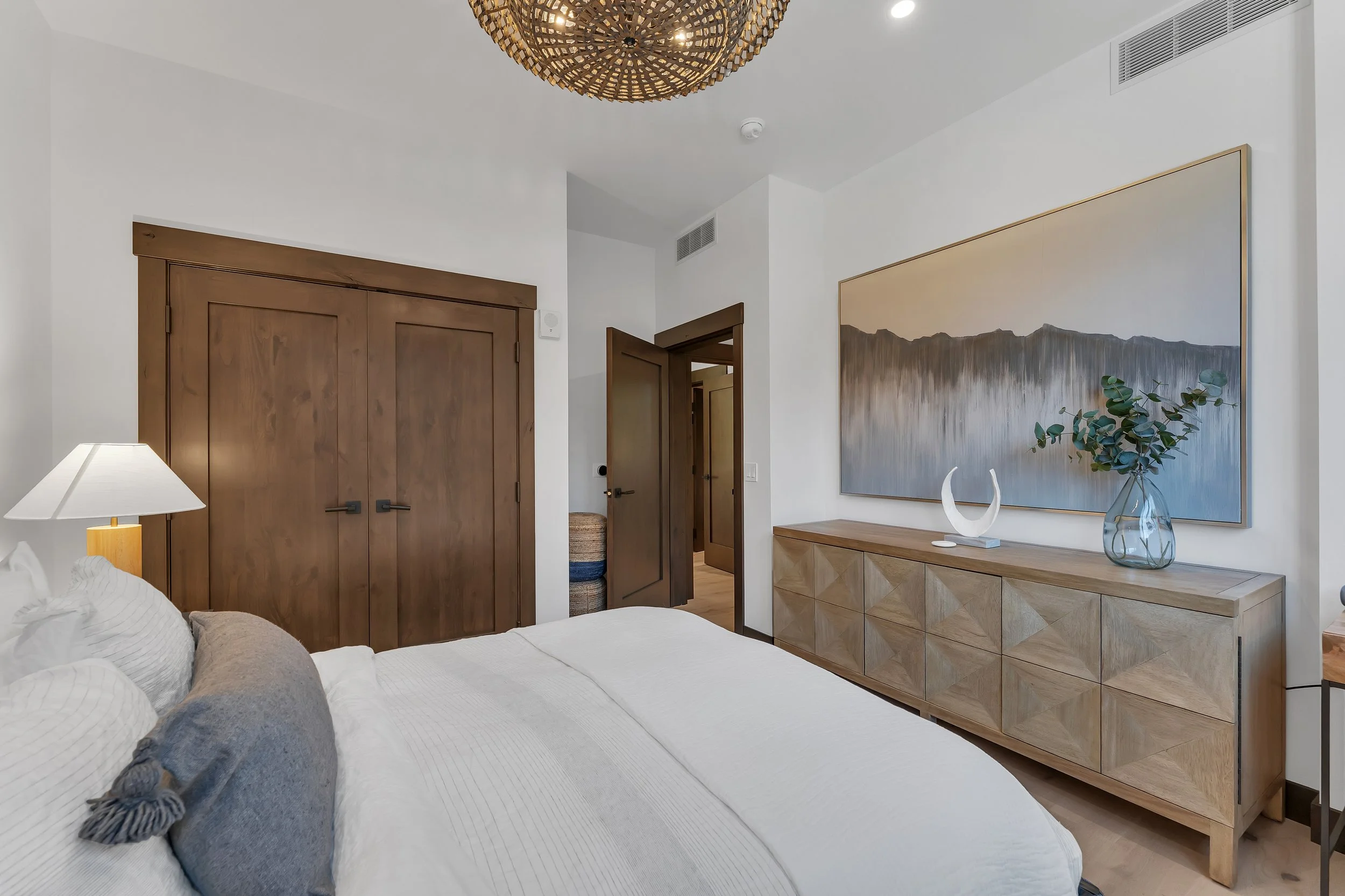 This cozy designer-furnished bedroom features a plush bed, a spacious light wood dresser, and thoughtfully placed artwork complemented by warm light hardwood floors.