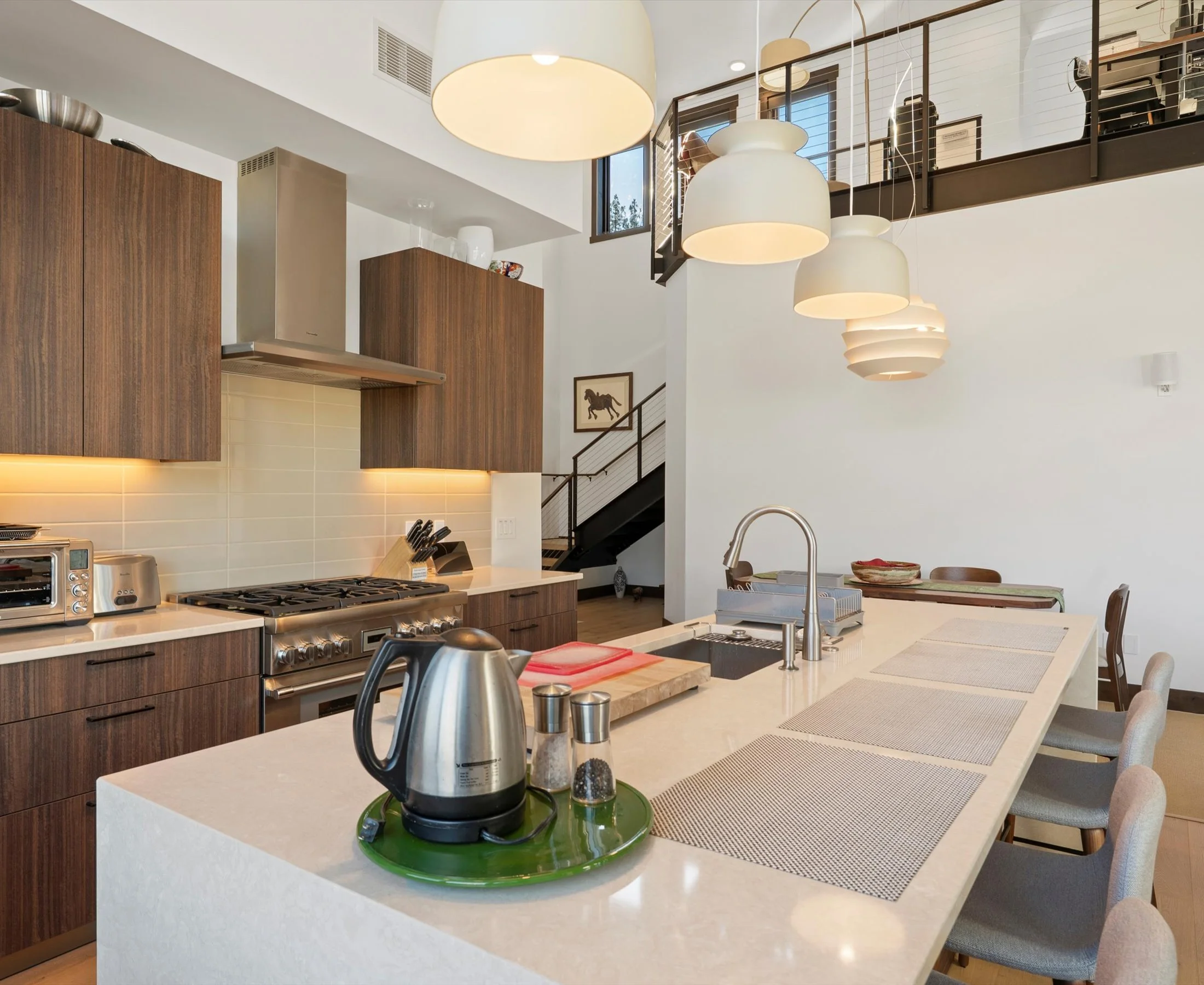 A pristine kitchen with a modern white island, barstools and stylish pendant lighting, offering a fresh and contemporary look. The Loft above is visible from the kitchen.