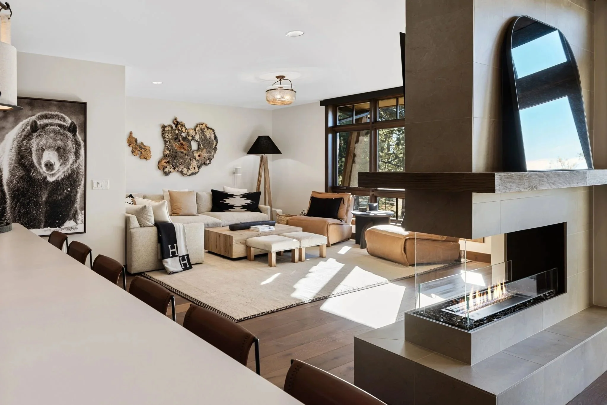 Open-concept luxury condo living room featuring a sleek double-sided gas fireplace, cream sectional sofa with Hermès throw, large-scale grizzly bear photography, organic wood wall art, and floor-to-ceiling windows with pine tree views