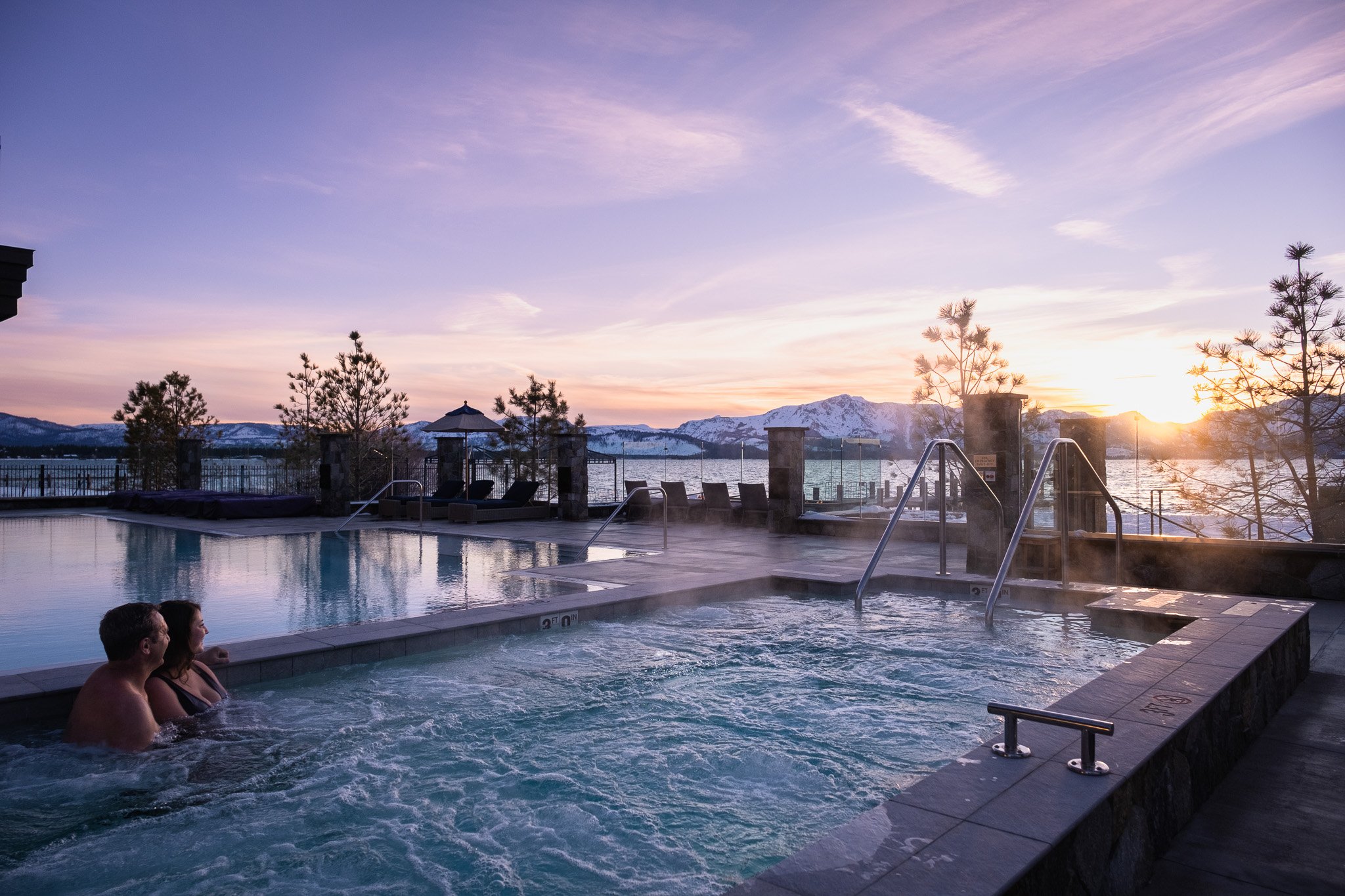 Two people sitting in a hot tub, basking in the soothing waters while the sun sets behind Lake Tahoe.
