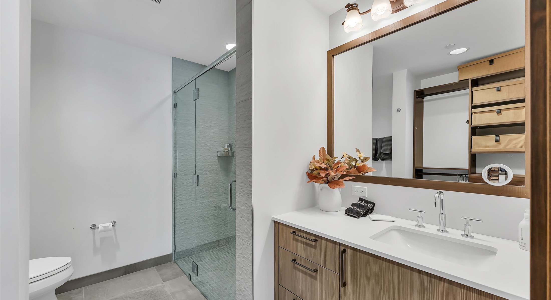 Modern and stylish bathroom featuring a a large vanity with wood cabinetry,  custom tiled floor, and a glass enclosed shower.