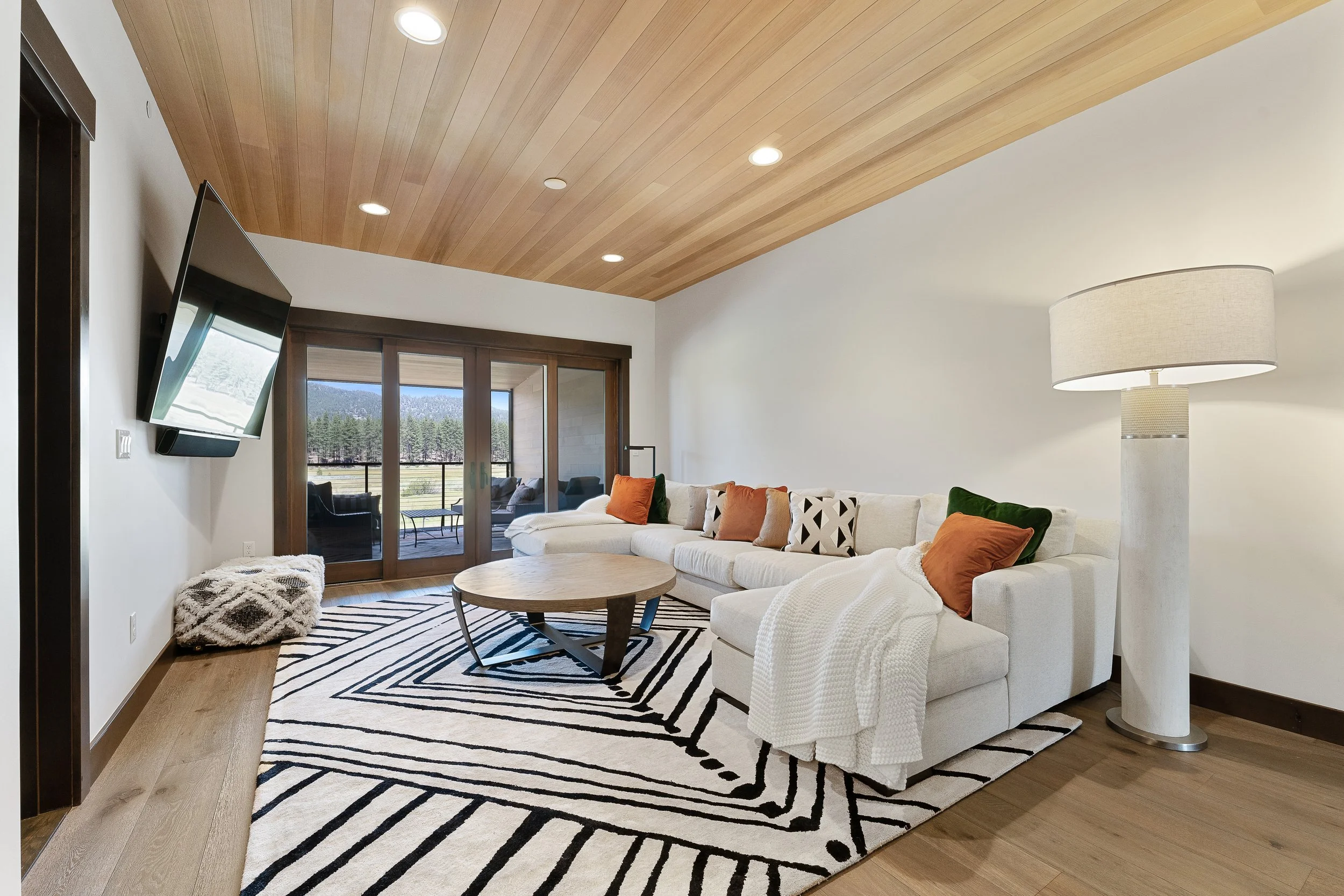 This cozy living space features natural wood ceiling, a large sofa with expansive windows framing serene forest views, and abundant natural light.