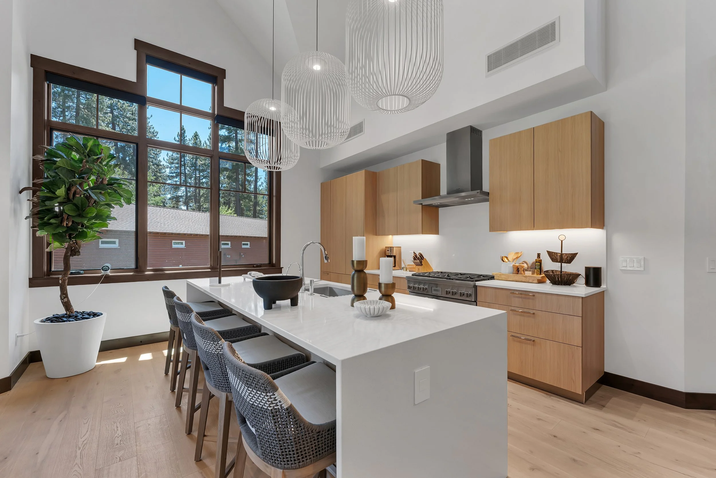 This open-concept kitchen is thoughtfully designer furnished and features light wood cabinetry, stainless steel appliances, hardwood floors, and a spacious white island with barstool seating.