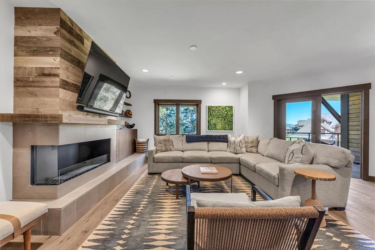 This cozy yet contemporary living area features a large seating area, warm stone fireplace, hardwood floors, an area rug and abundant natural light streaming through multiple windows. A patio door opens seamlessly to a private outdoor retreat.