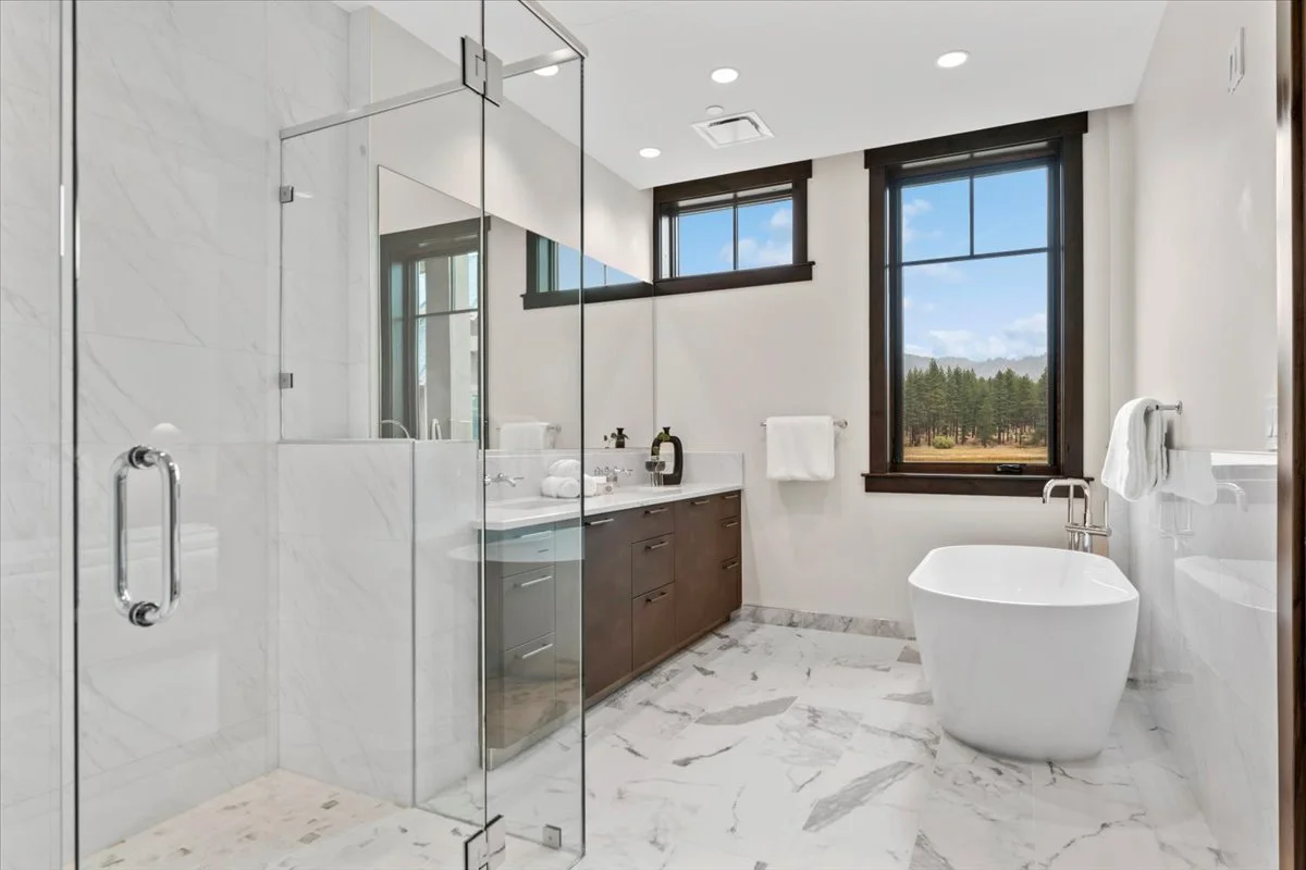 Bright and sophisticated bathroom featuring custom tile, a glass-enclosed shower, modern freestanding tub, dual vanity cabinetry, and tranquil forest views.