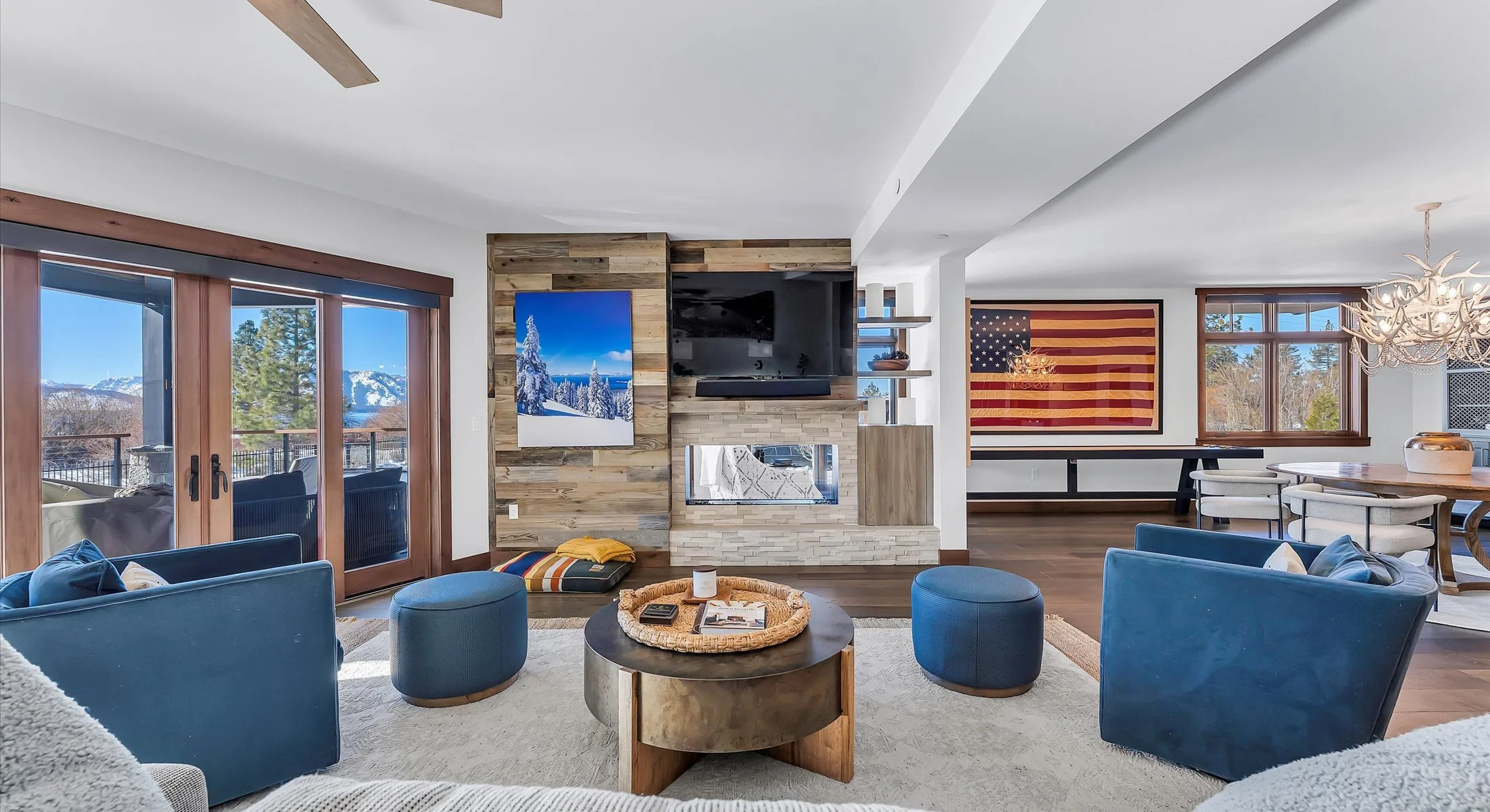 This stylish open-concept living area blends comfort and sophistication, featuring wide-plank hardwood floors, a cozy dual-sided stone fireplace with rustic accents, and 2 large 4-panel glass doors leading out to a private patio while letting in an a