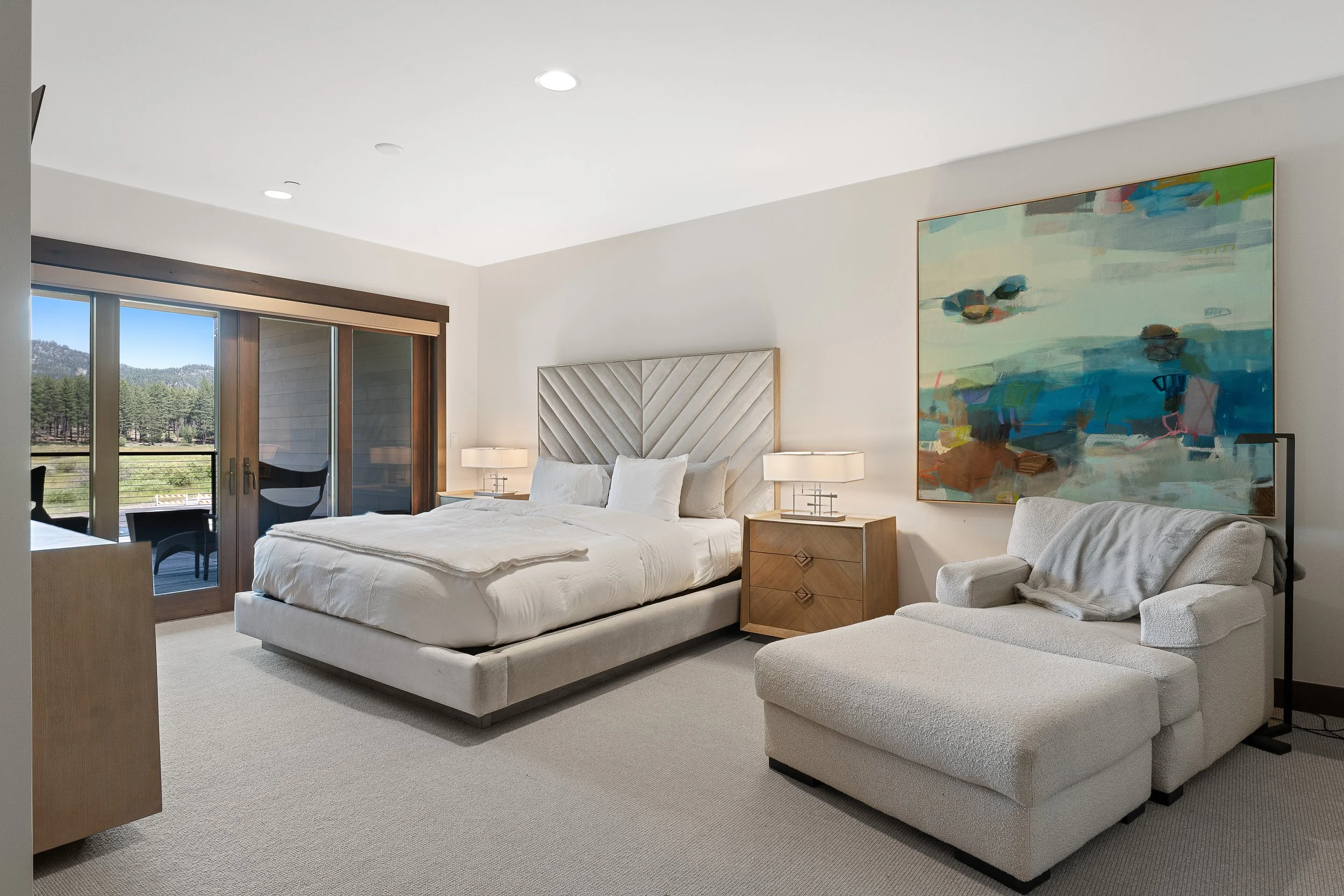 Contemporary bedroom featuring a large bed, bright carpet, sitting area perfect for reading and sophisticated design accents, with a door opening to a private patio overlooking the serene forest.