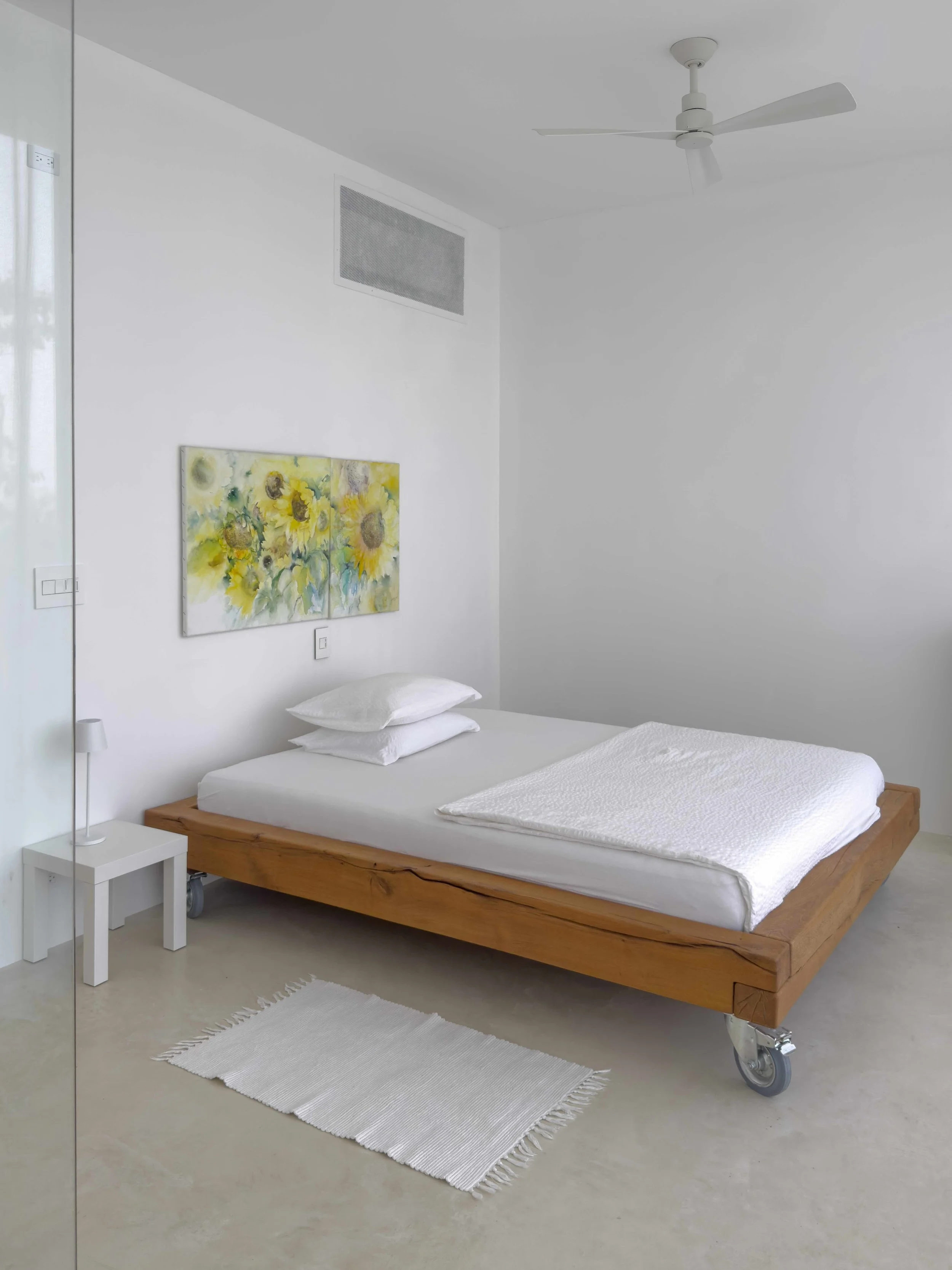 Minimalist bedroom with a wooden bed on wheels, white bedding, a small white bedside table with a lamp, a white rug, sunflower painting on white wall, ceiling fan, and vent on ceiling.