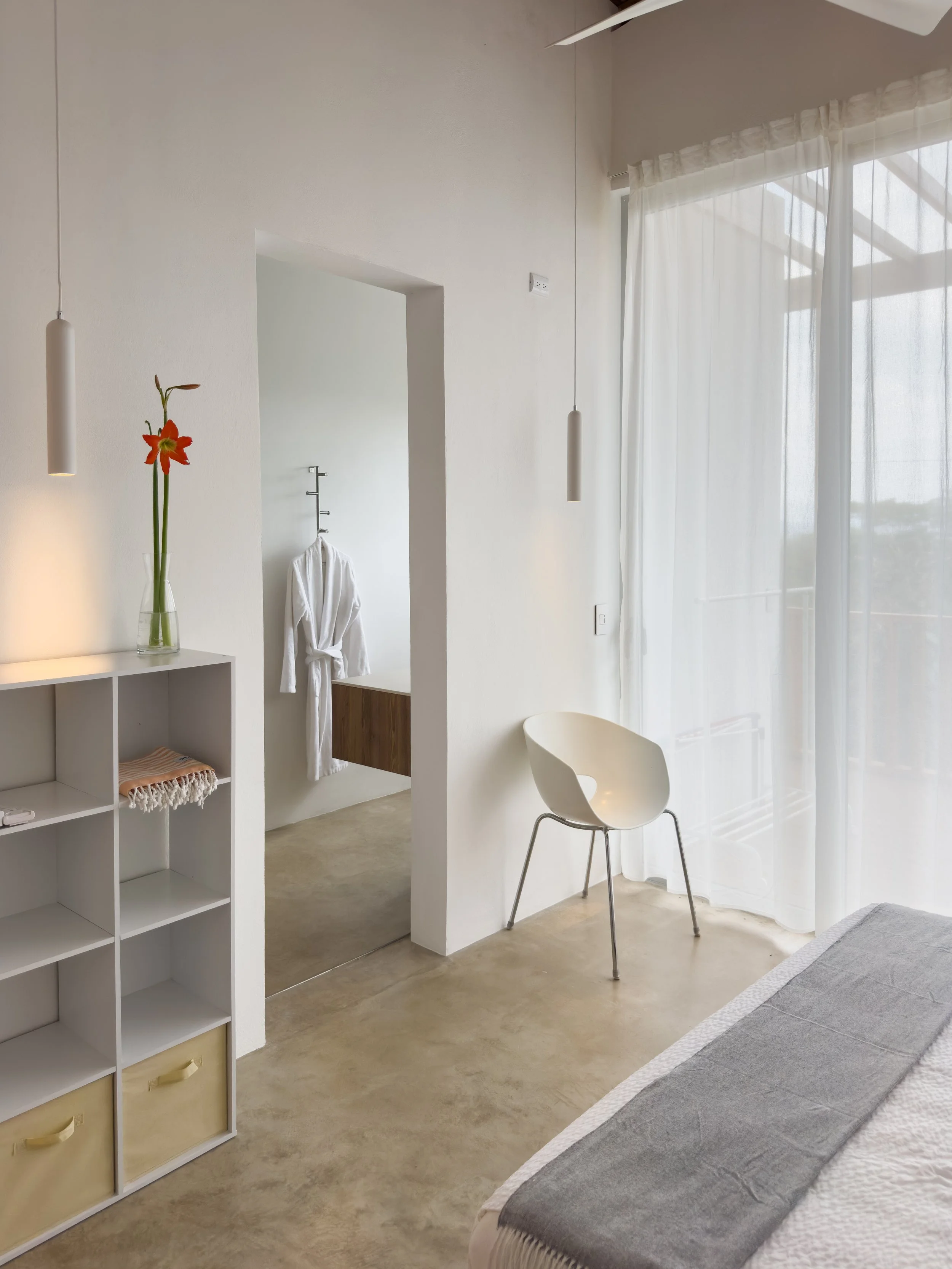 A minimalist bedroom with white walls, a bedside table with a glass vase and red flower, a white chair, a bed with gray and white bedding, and sheer curtains letting in natural light.