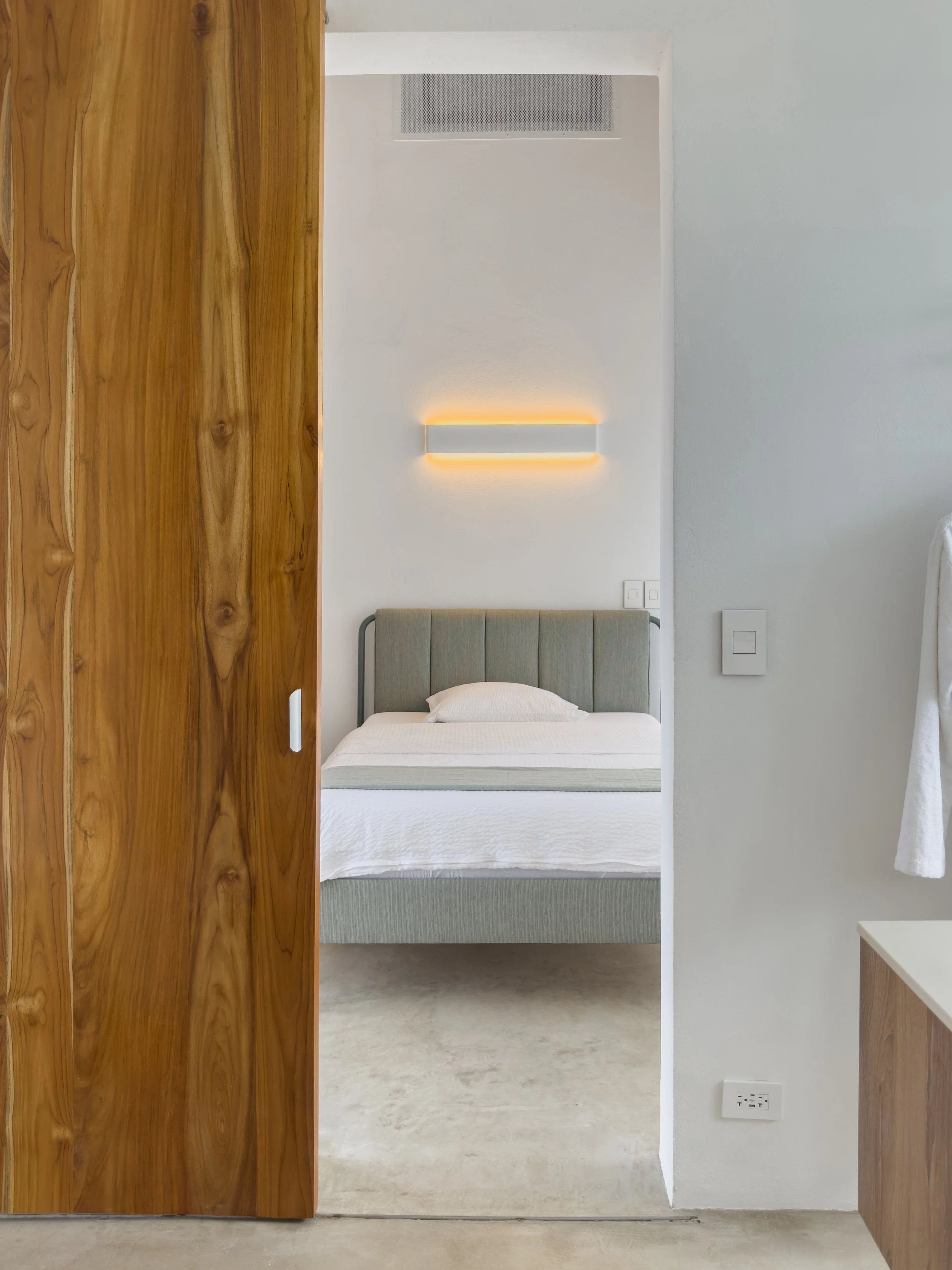 View of a minimalist bedroom with a light green upholstered bed, a white wall-mounted light fixture, and a partially opened wooden sliding door revealing the room's interior